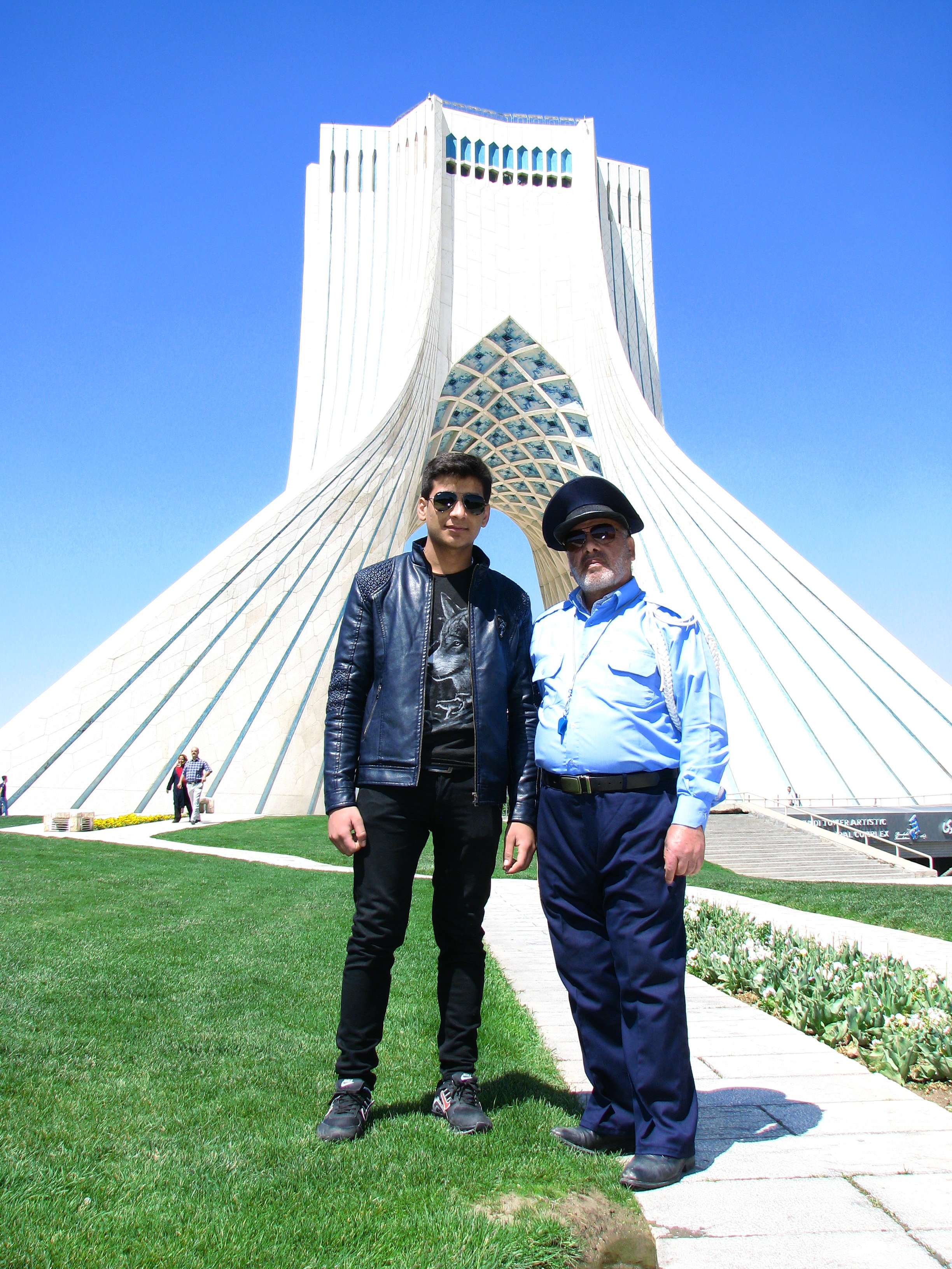 Two men standing on grass in front of Azadi Tower in Tehran, Iran, with blue sky background.
