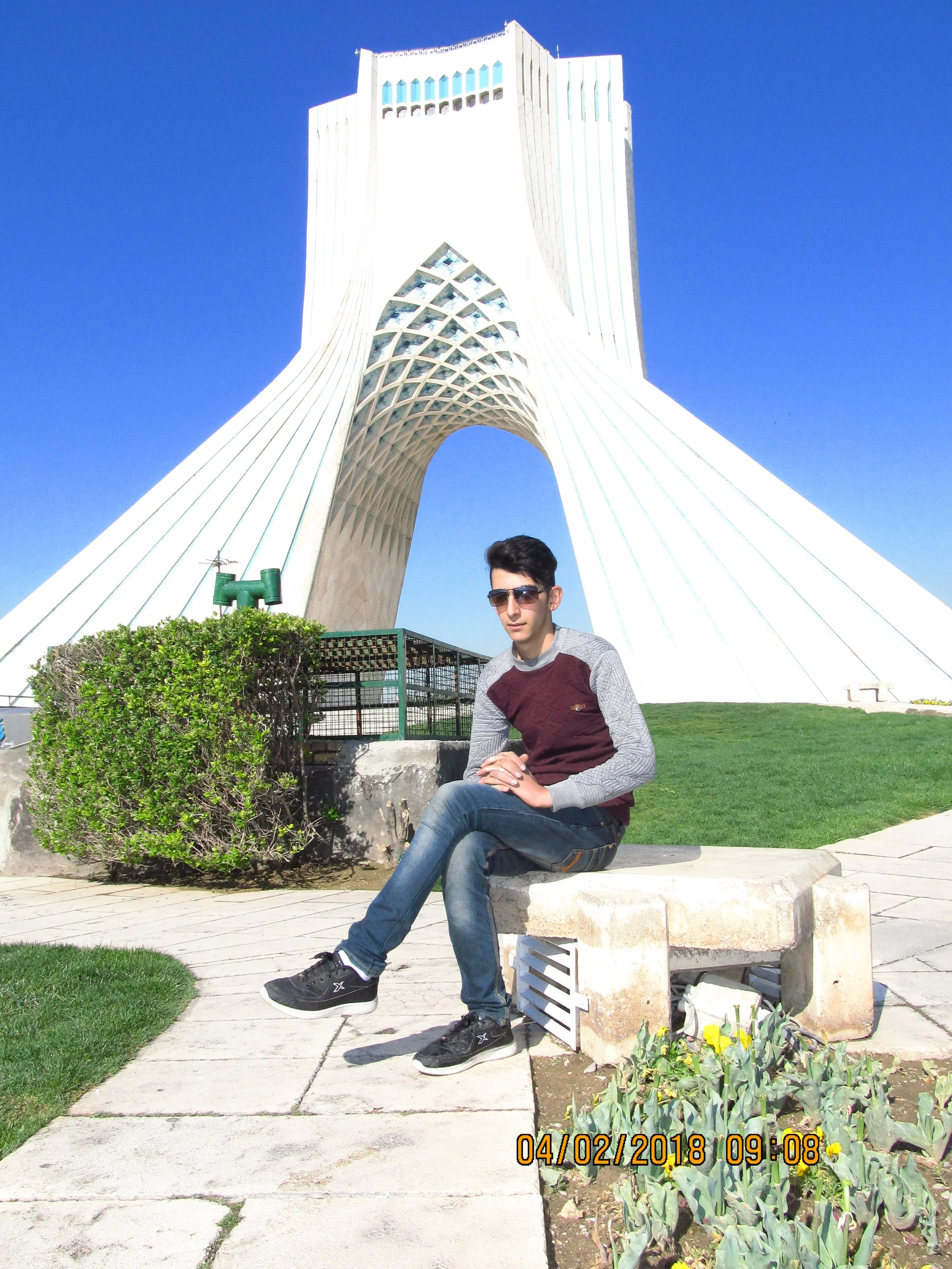 Young man sitting on a stone bench outdoors in front of a large architectural structure with a blue sky background.