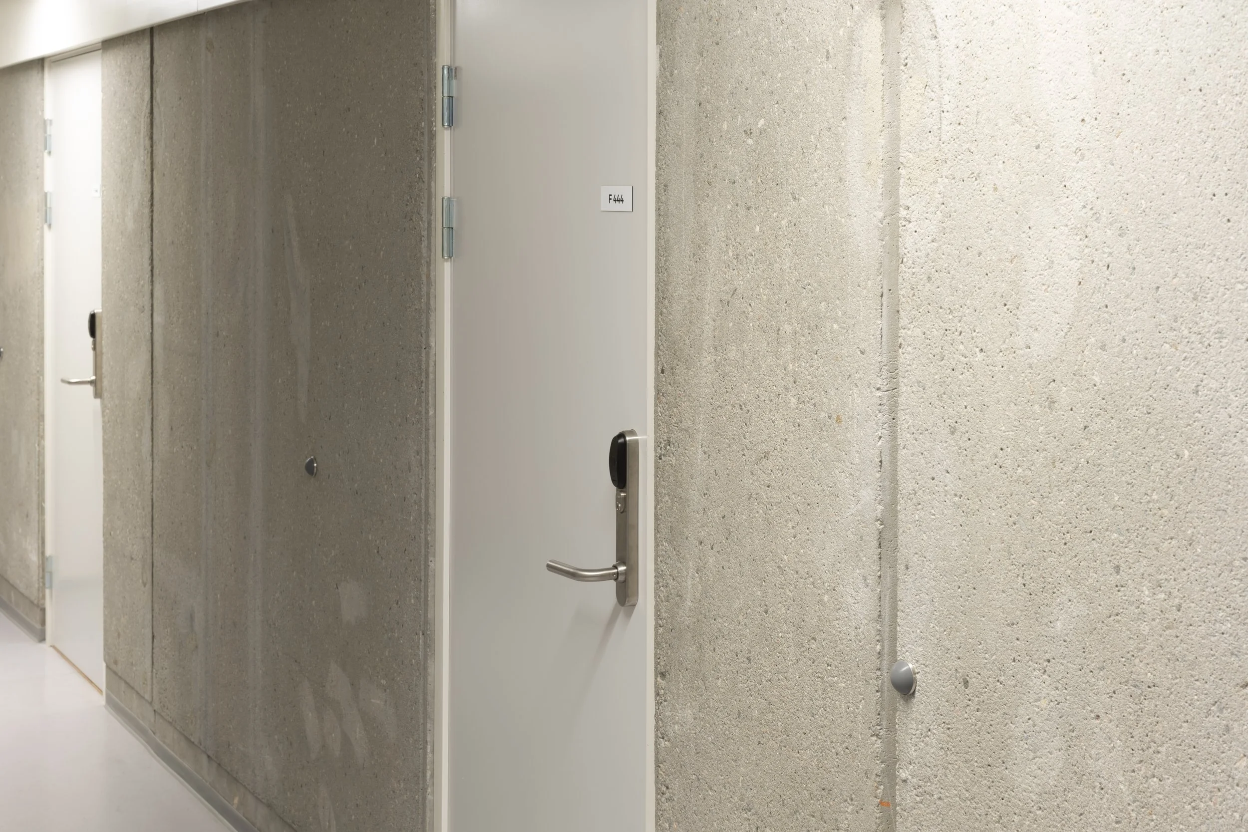 A hallway with concrete walls and white doors, one door has a black lock and a small label reading 'F444'.