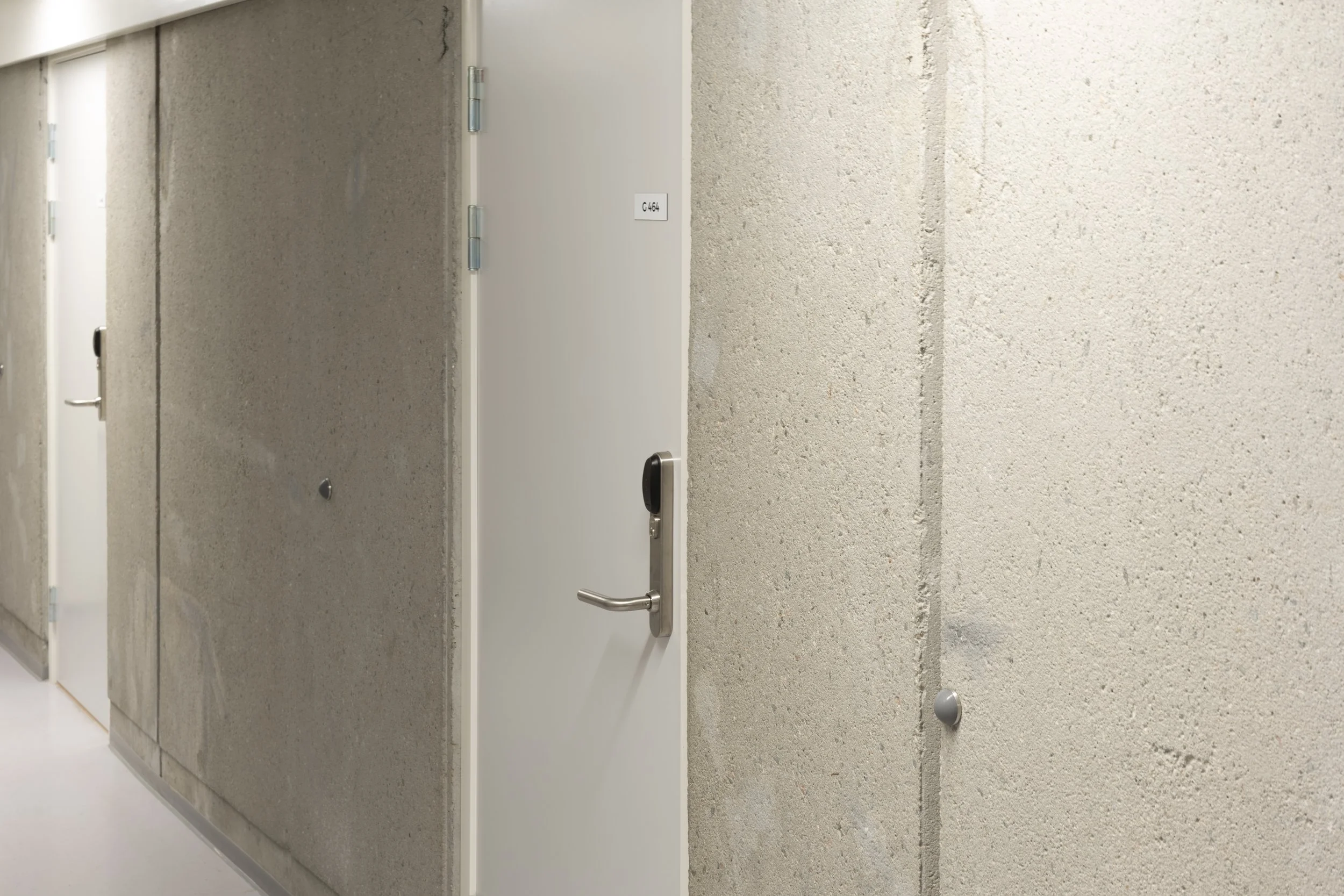 A corridor with the brown and beige wall on the right side and a row of white doors on the left, two of which have their doors open.