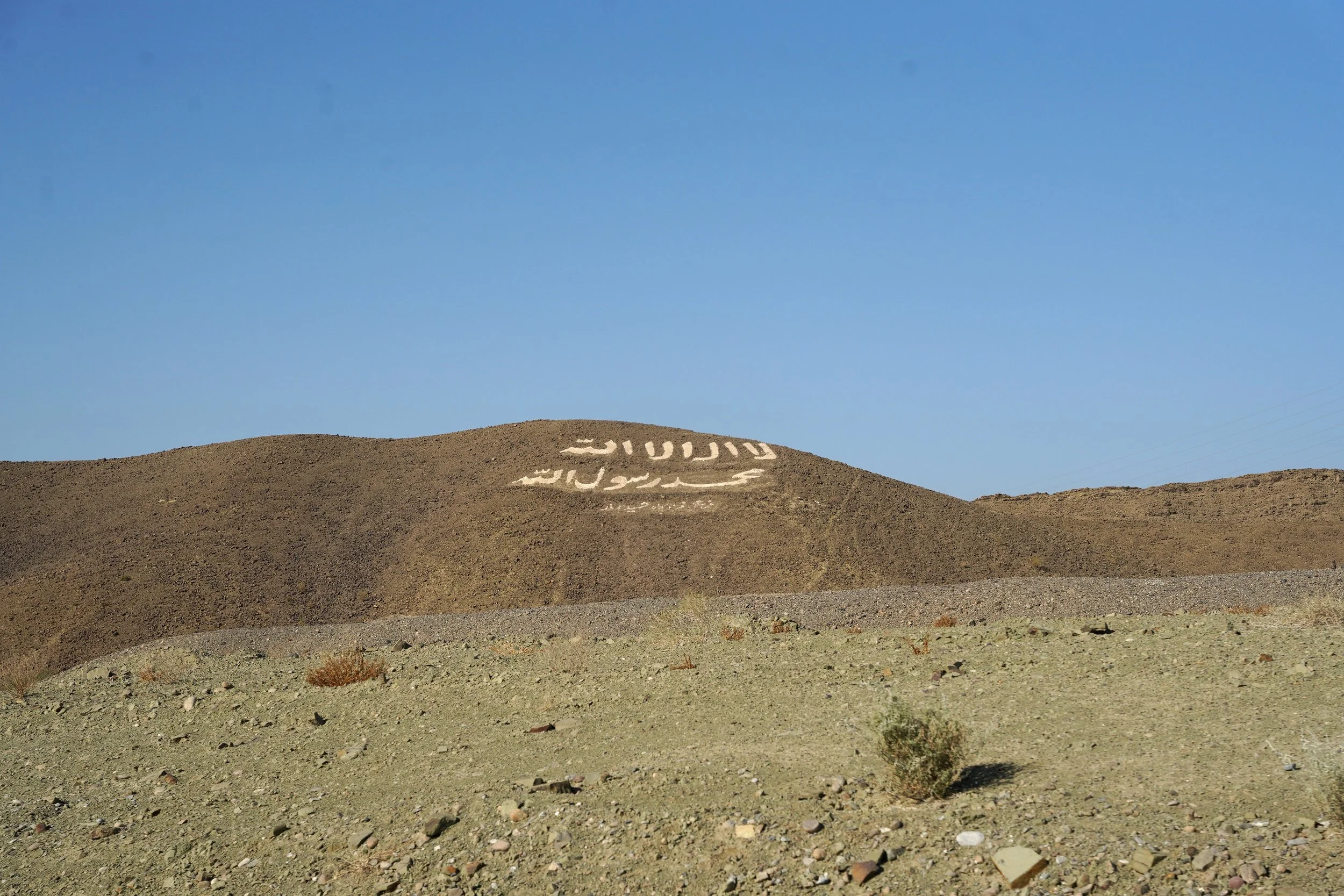 Arabic script in white on brown hillside under clear blue sky.
