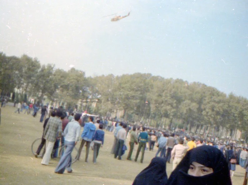 Crowd of people standing in an open field with a helicopter flying overhead, trees in the background, and partially visible woman wearing a black hijab in the foreground.