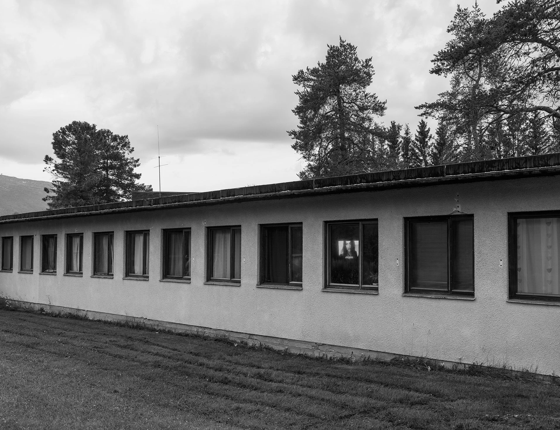 A black and white photo of a single-story building with multiple windows, some of which have curtains, and a reflection of a woman's face in one window. There are tall trees and cloudy sky in the background.