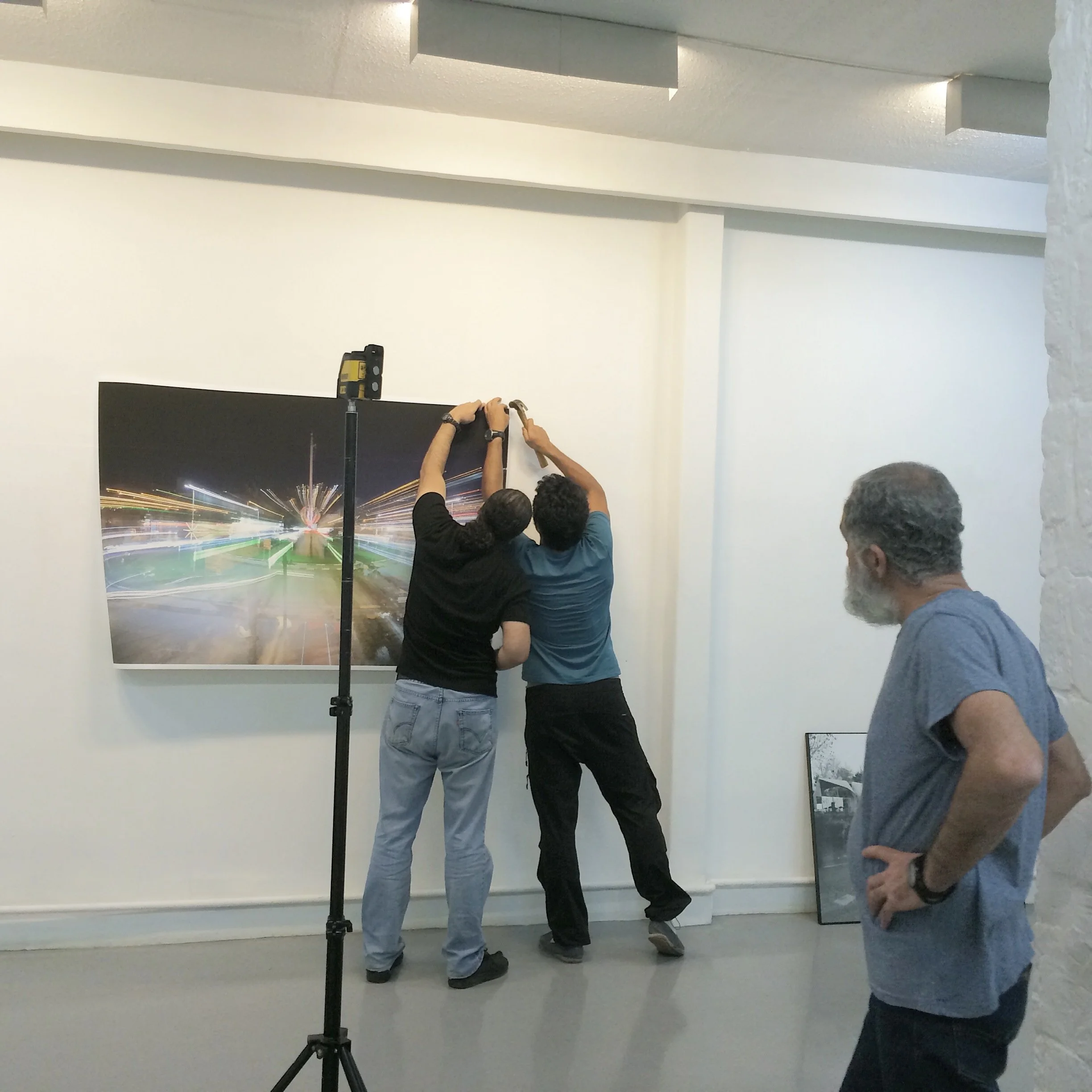 Two men are hanging a large photograph or artwork on a white gallery wall, while a third man observes with his hands on his hips in an art gallery or exhibition space.