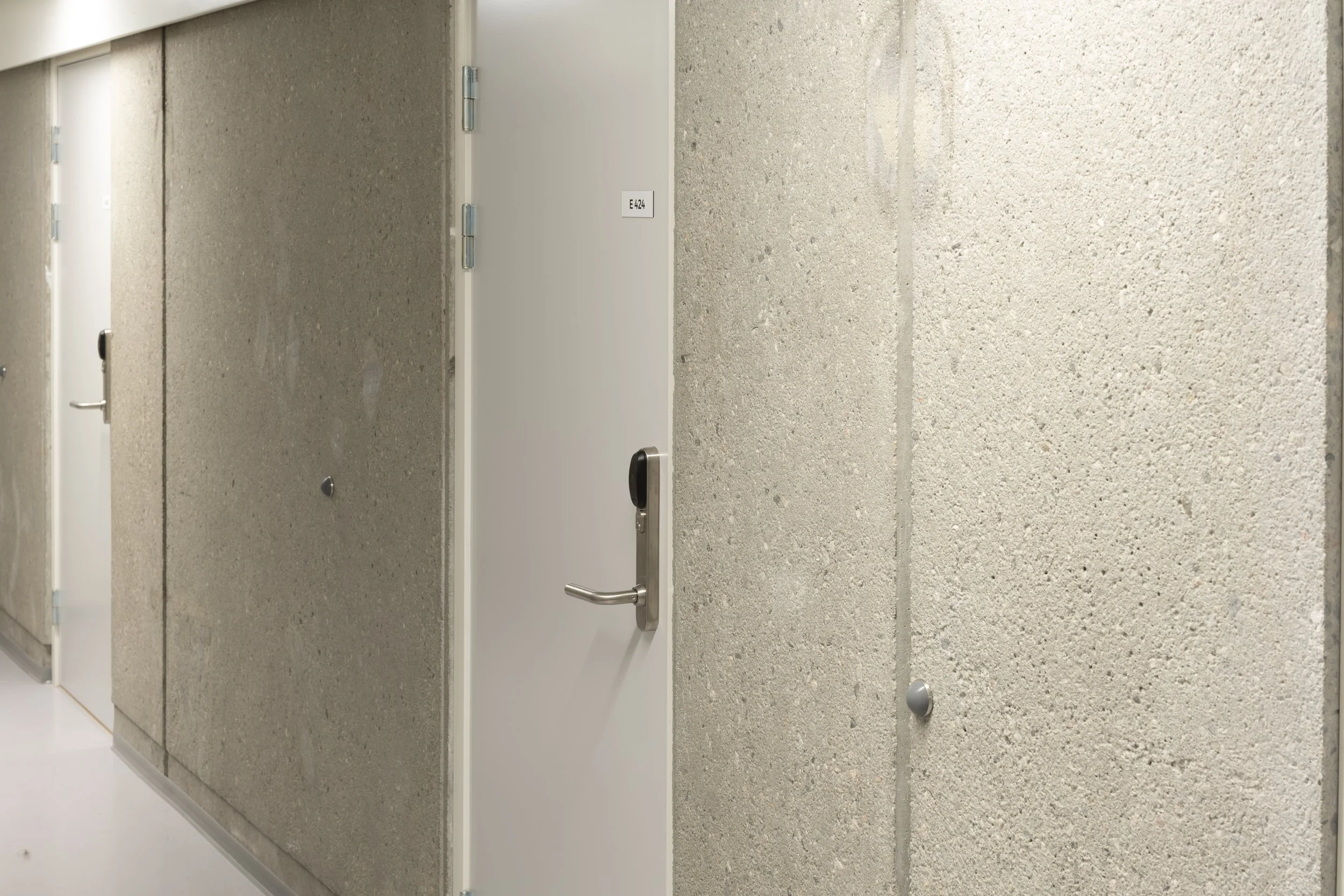Hallway with plain concrete walls, white doors, and metallic handles, some doors labeled with room numbers.