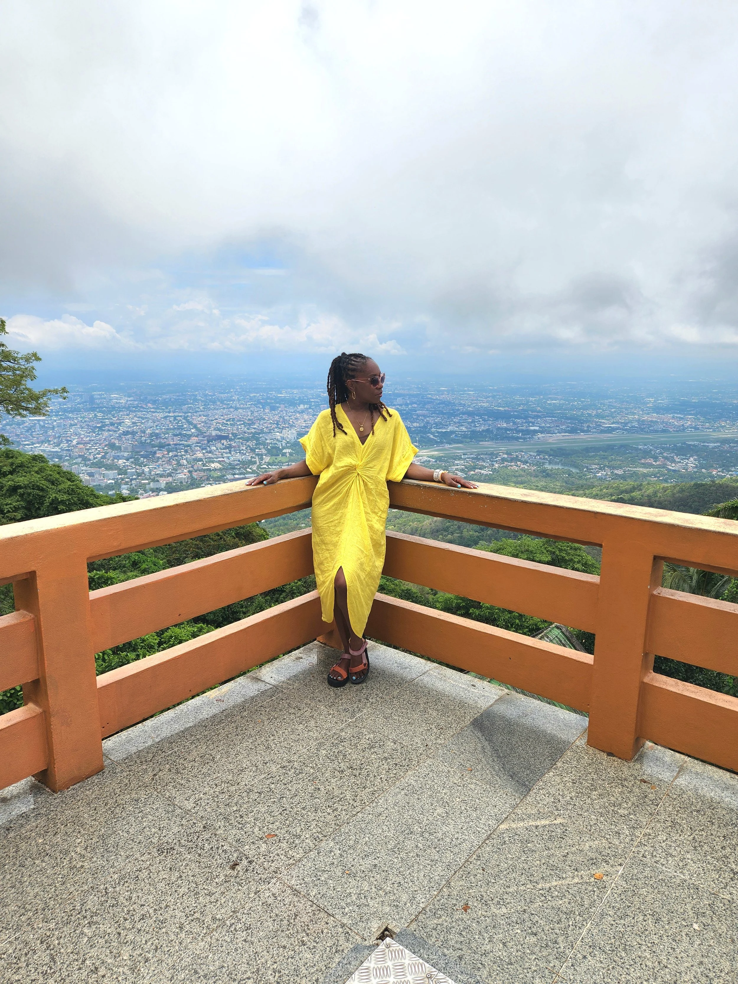 A woman in a yellow dress stands at a viewing point with a cityscape and cloudy sky behind her, leaning on a wooden railing.