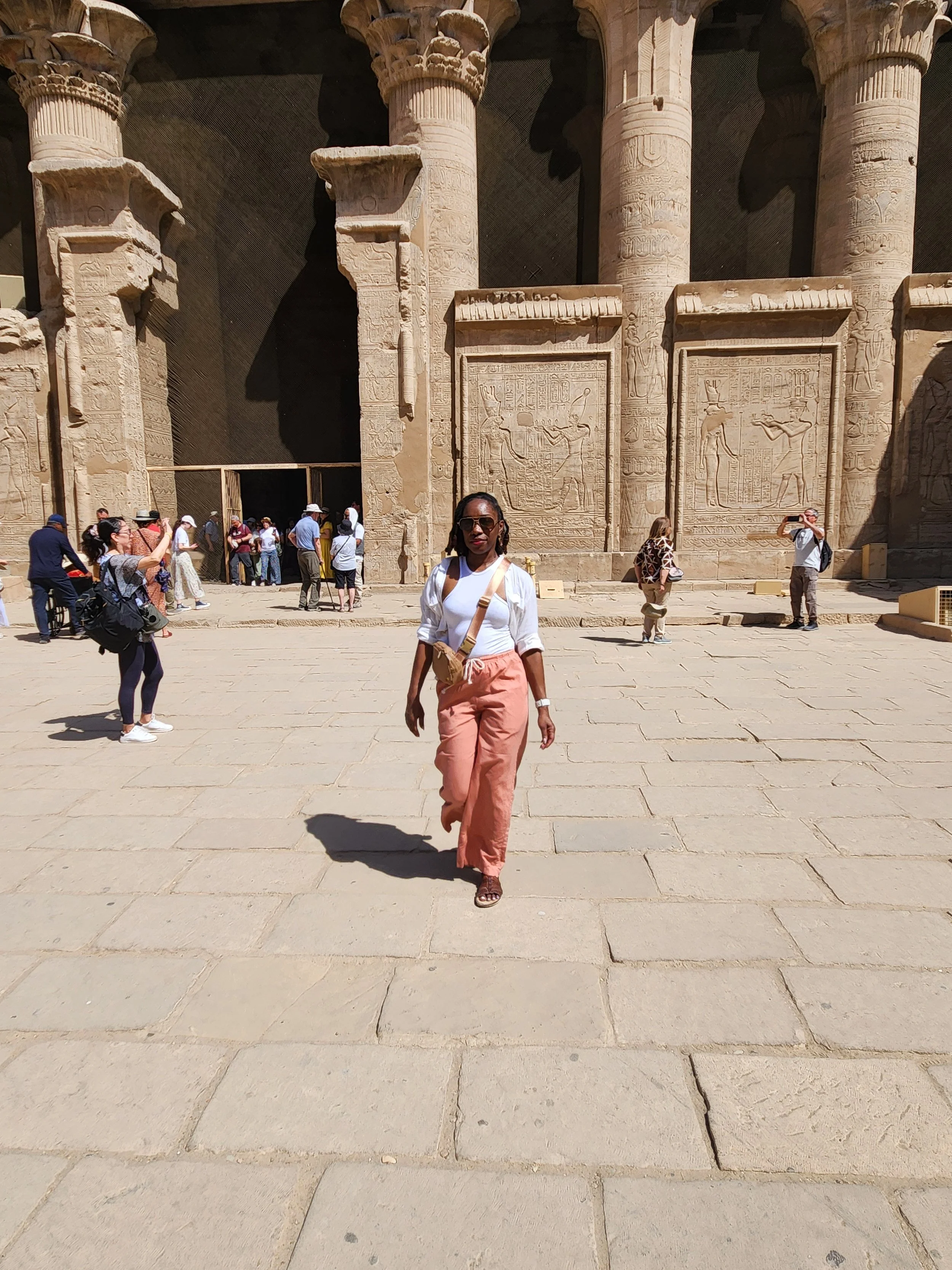A woman with sunglasses wearing a white top and orange pants walking in front of ancient Egyptian temple walls with hieroglyphics and carvings, surrounded by other tourists.