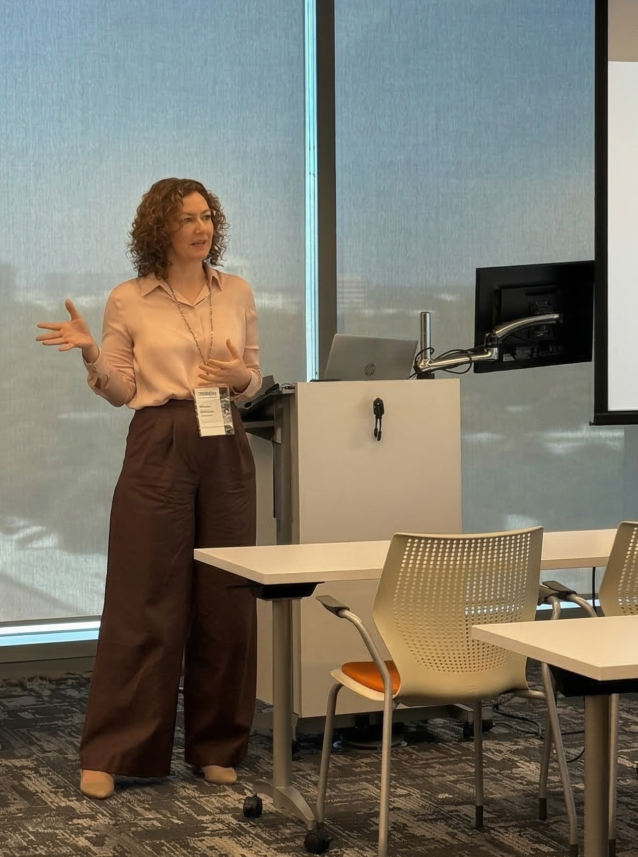 A woman with curly hair giving a presentation in a conference room, standing next to a podium with a laptop and a monitor. She is wearing a light pink blouse and dark brown pants.