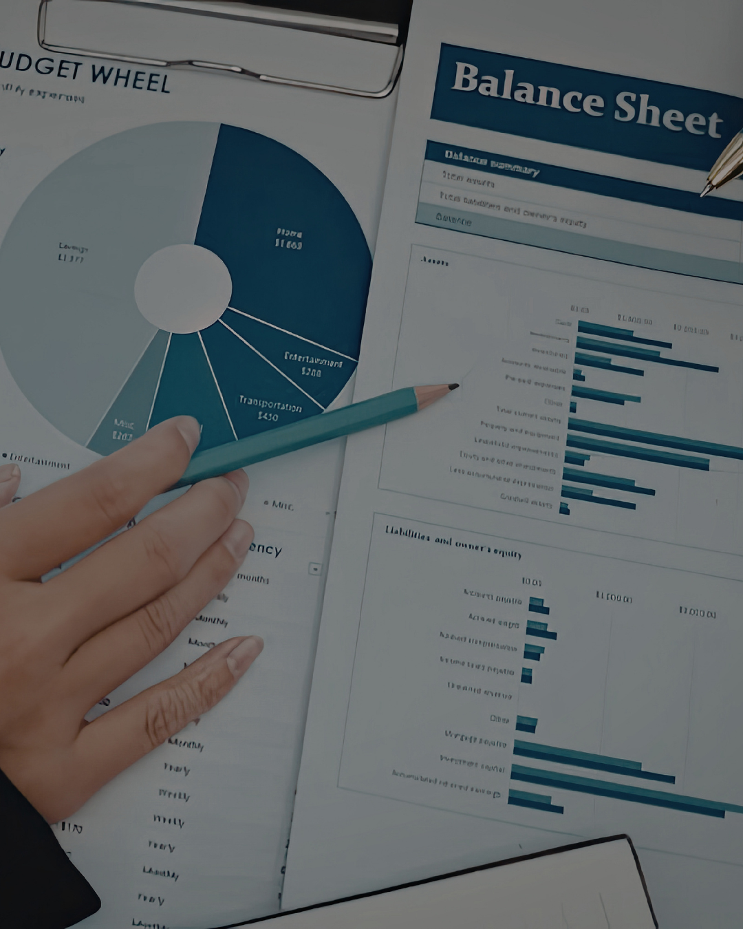 Hand holding a blue pencil pointing at a financial report with bar charts and a balance sheet on a desk, surrounded by documents and charts.