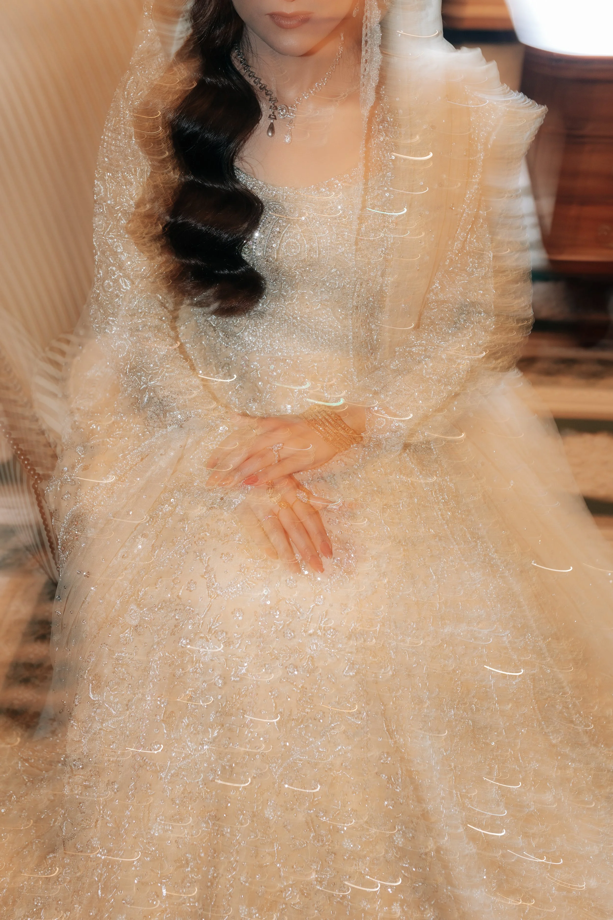 Woman dressed in an elaborate, shimmering beige or champagne gown with lace and bead details, wearing jewelry including a necklace and rings, with a veil on her head, possibly a bride in a wedding setting.