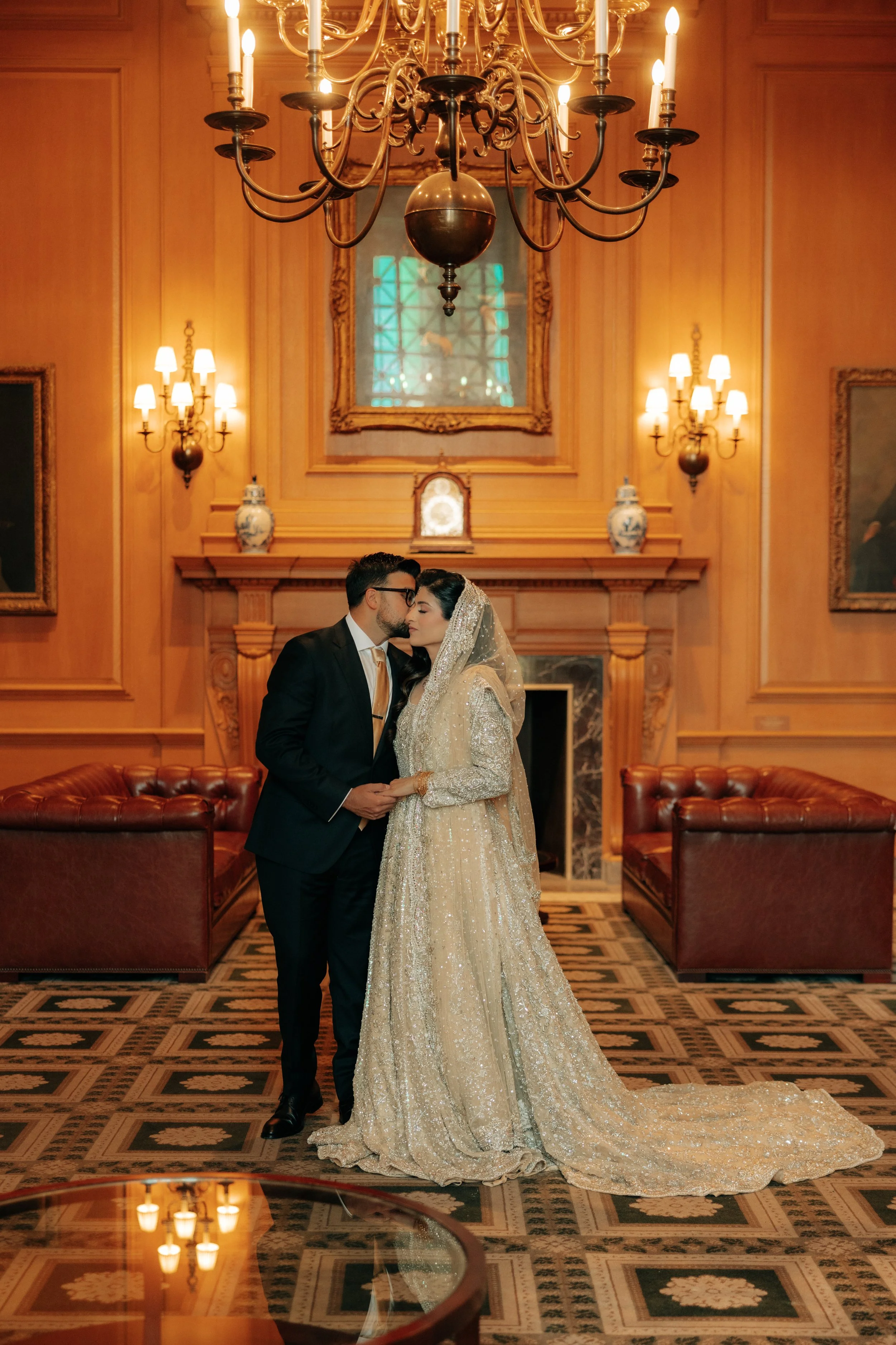 A bride and groom sharing a close moment in an elegant, warmly lit room with a large chandelier, ornate mirror, framed artwork, and vintage-style furniture.