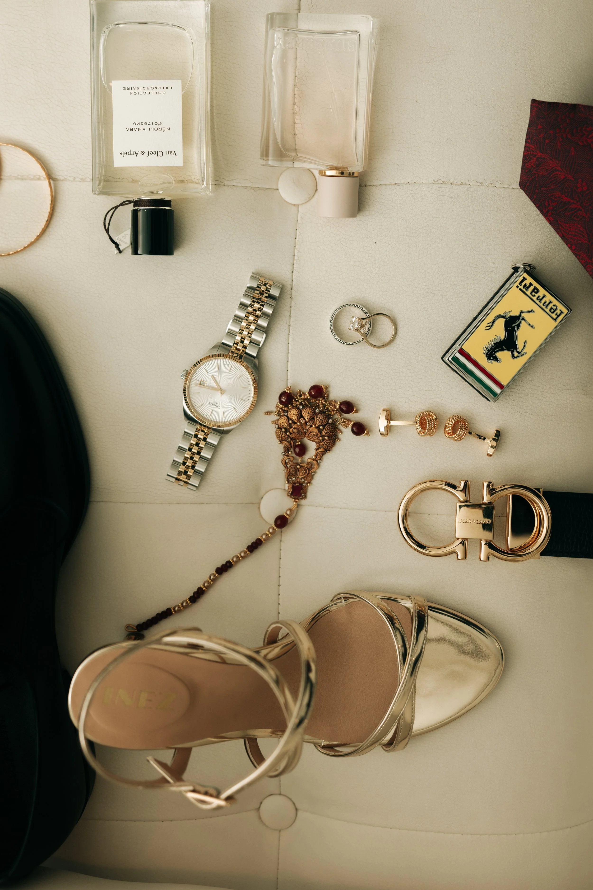 Collection of jewelry and accessories on a beige surface, including a gold and silver watch, a gold necklace with red stones, matching earrings, a gold ring, a gold bracelet, a pair of pink high-heeled shoes, a rectangular fashion lighter with a hors
