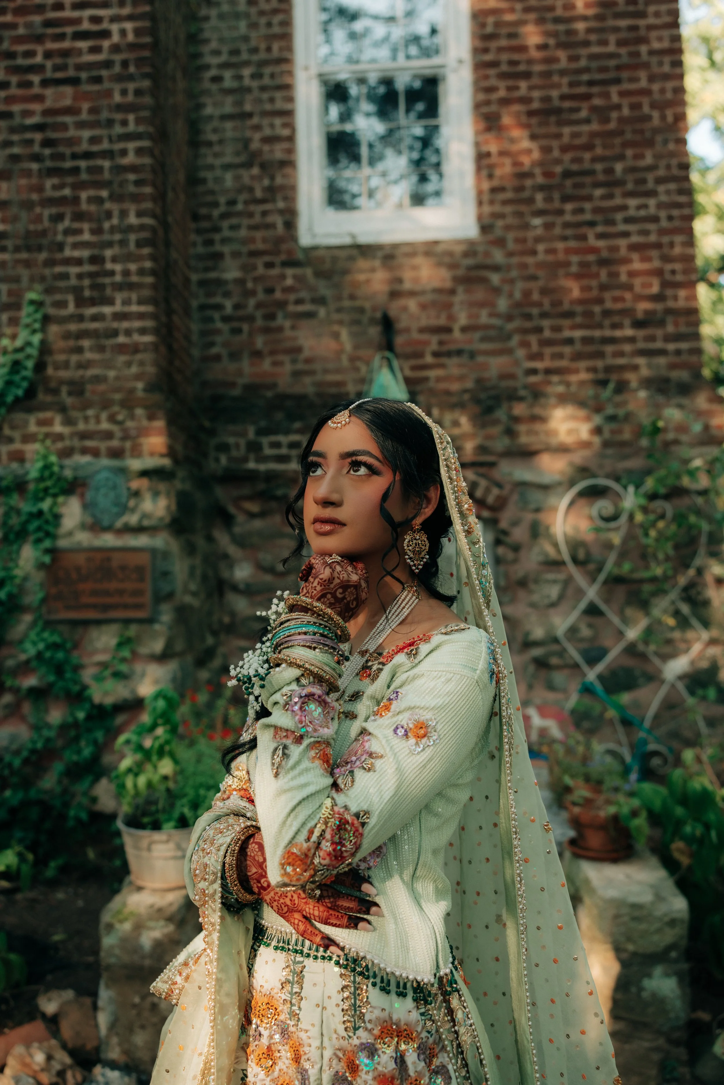 Young woman wearing traditional South Asian bridal attire with ornate jewelry, bangles, and embroidery, standing outdoors in front of a brick wall with a window and greenery.