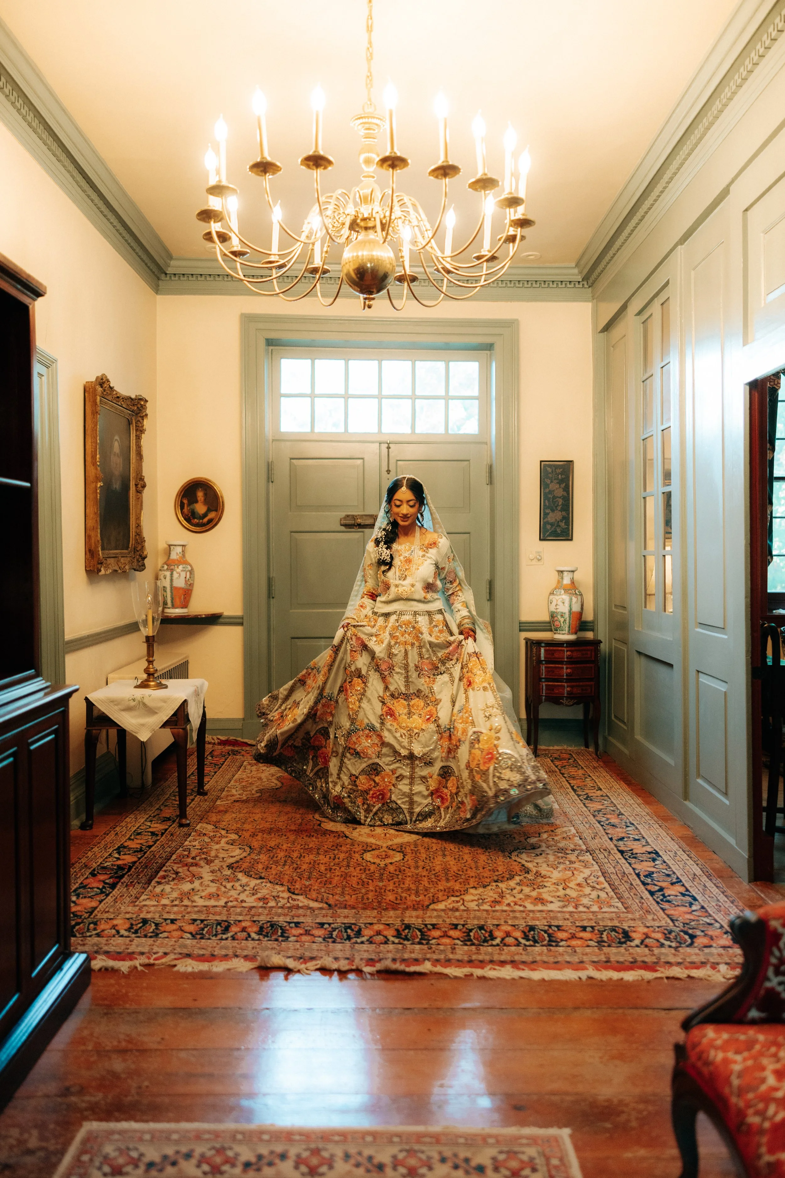 A woman in a traditional, ornate dress standing in a decorated living room with classic furniture and a large chandelier hanging from the ceiling.