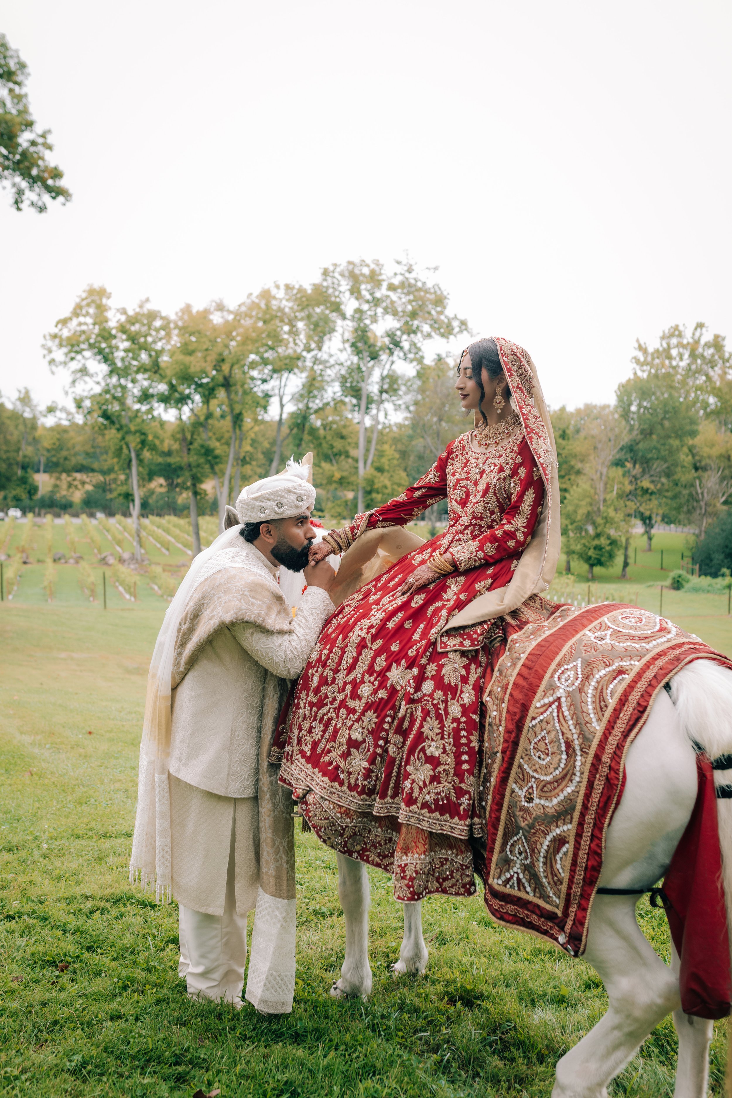 A bride in a red, embroidered wedding dress and traditional jewelry sits on a decorated horse, with a groom in cream traditional attire kneeling and kissing her foot, in a green outdoor setting with trees in the background.