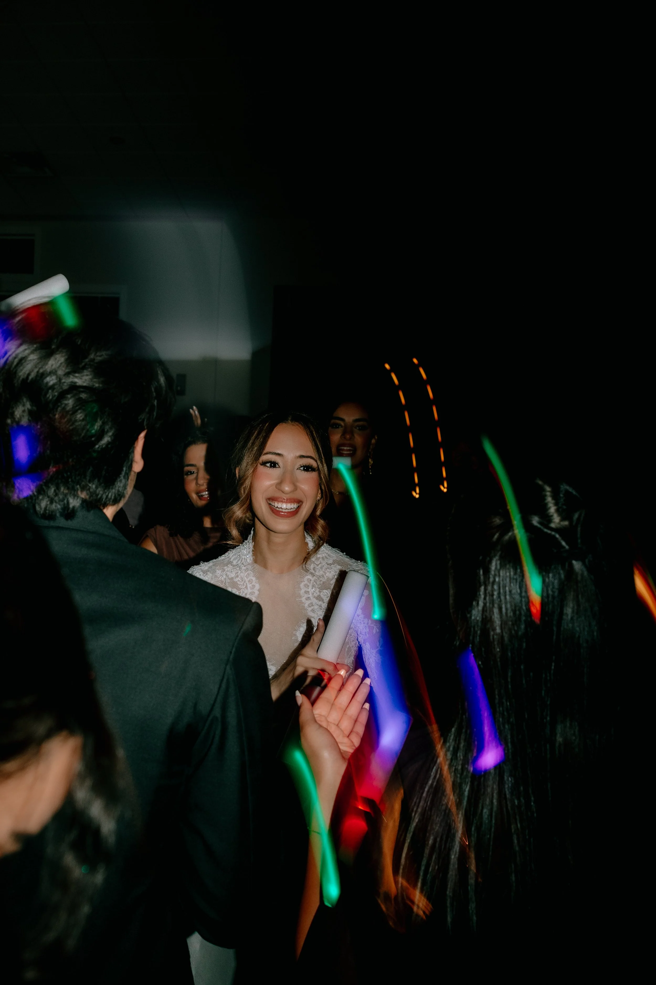 A group of partygoers celebrating, with a woman smiling in the center holding a glow stick, colorful light trails, and individuals in the background.