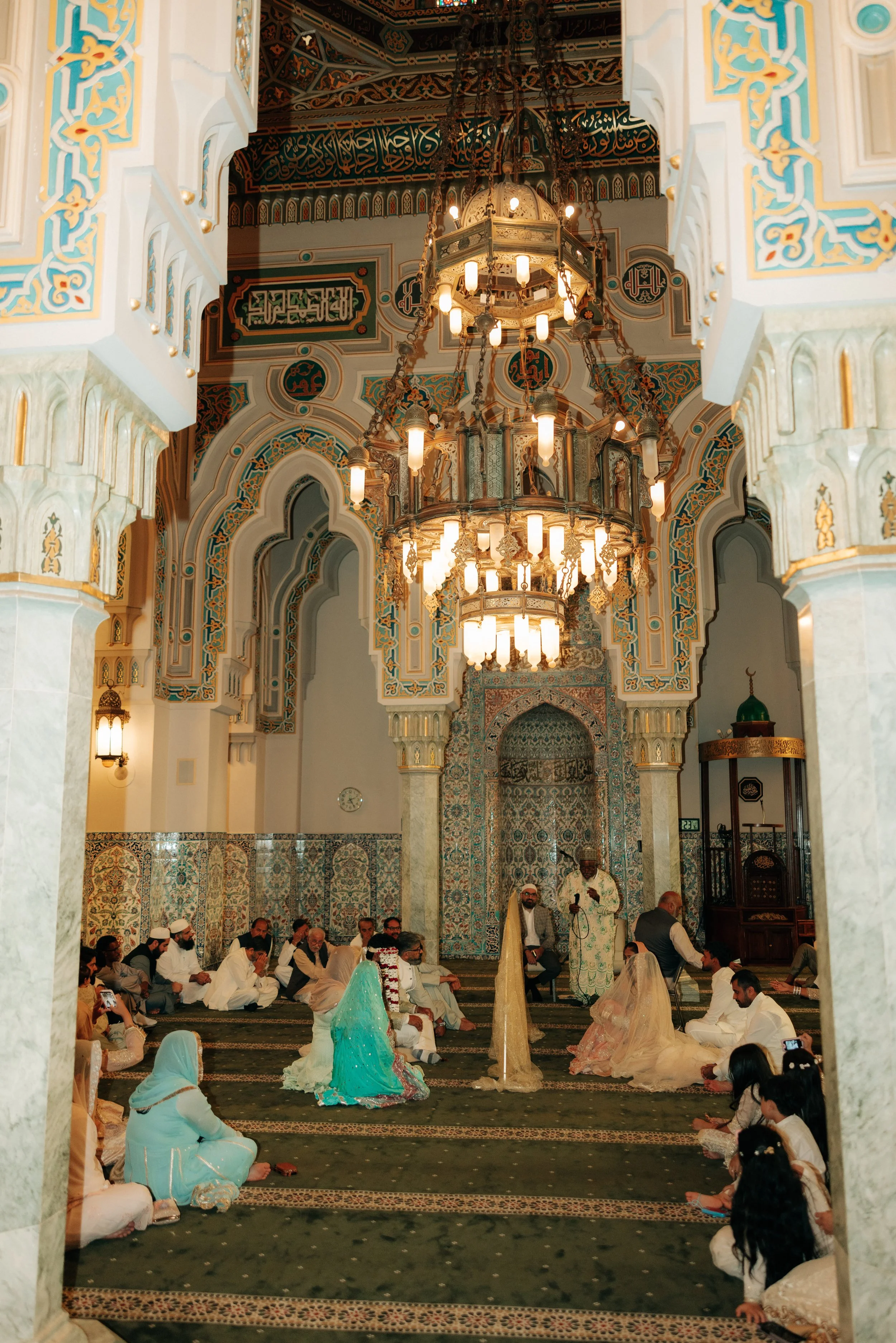 People seated on the floor inside a mosque, participating in a religious gathering or prayer. The mosque features ornate chandeliers, intricate wall and ceiling designs, and a mihrab at the front.