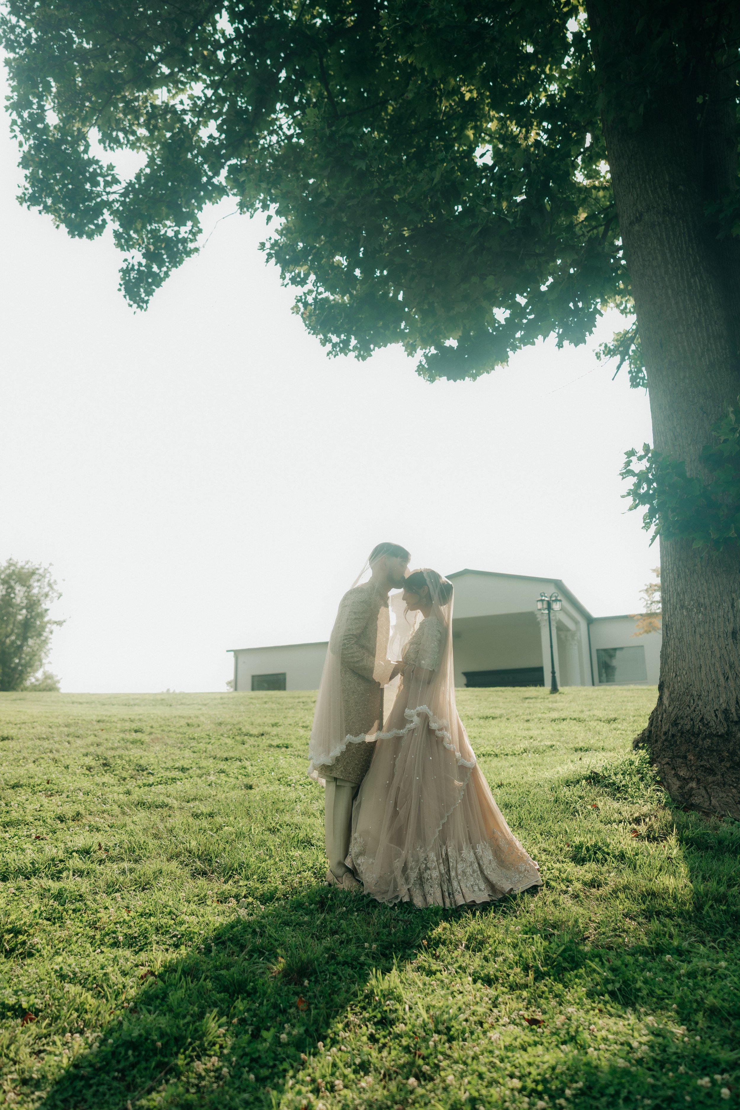 A bride and groom standing on a grassy lawn, holding hands and touching foreheads, with large trees and a building in the background.