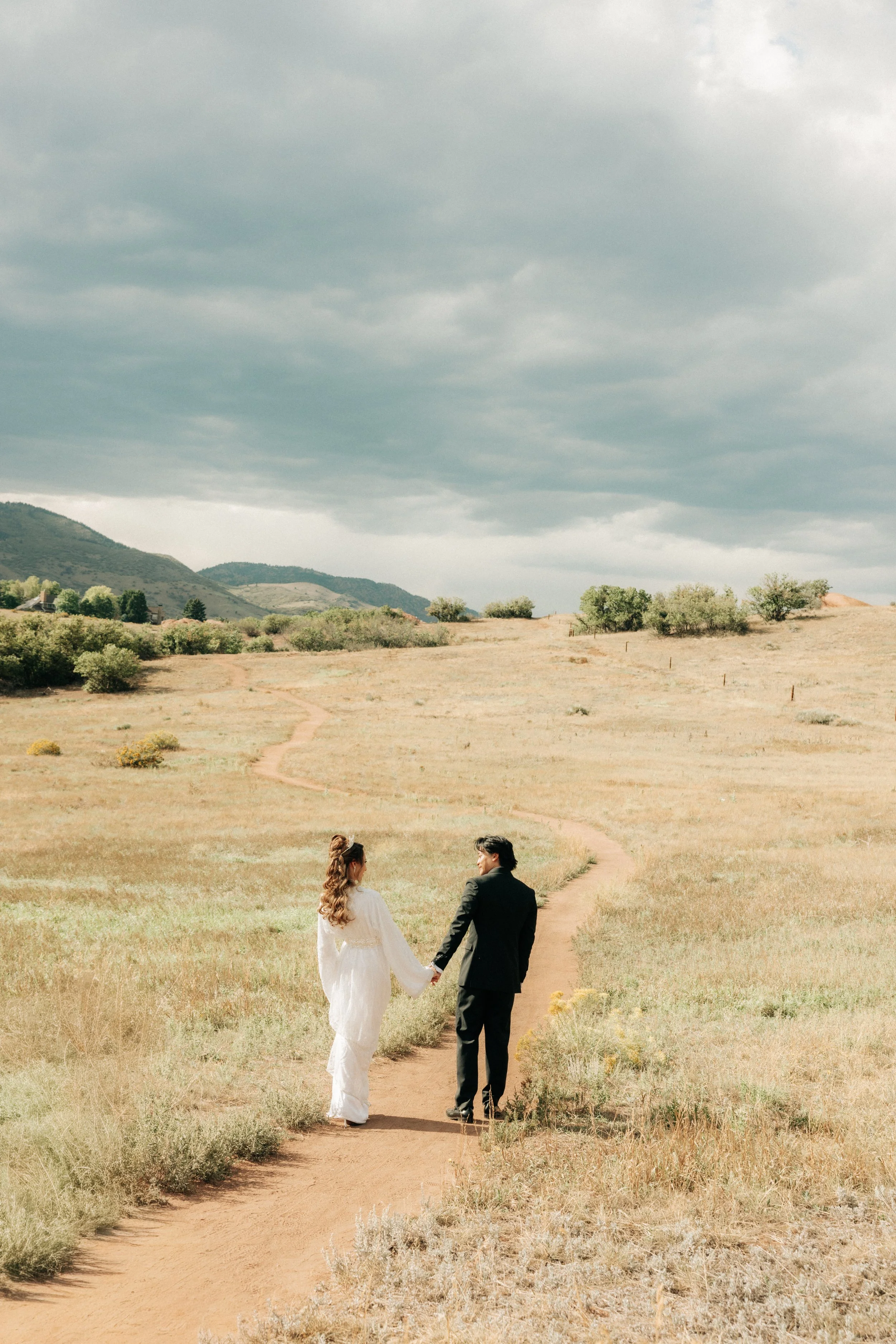 A bride and groom holding hands and walking along a dirt path in a grassy field, with hills and cloudy sky in the background.