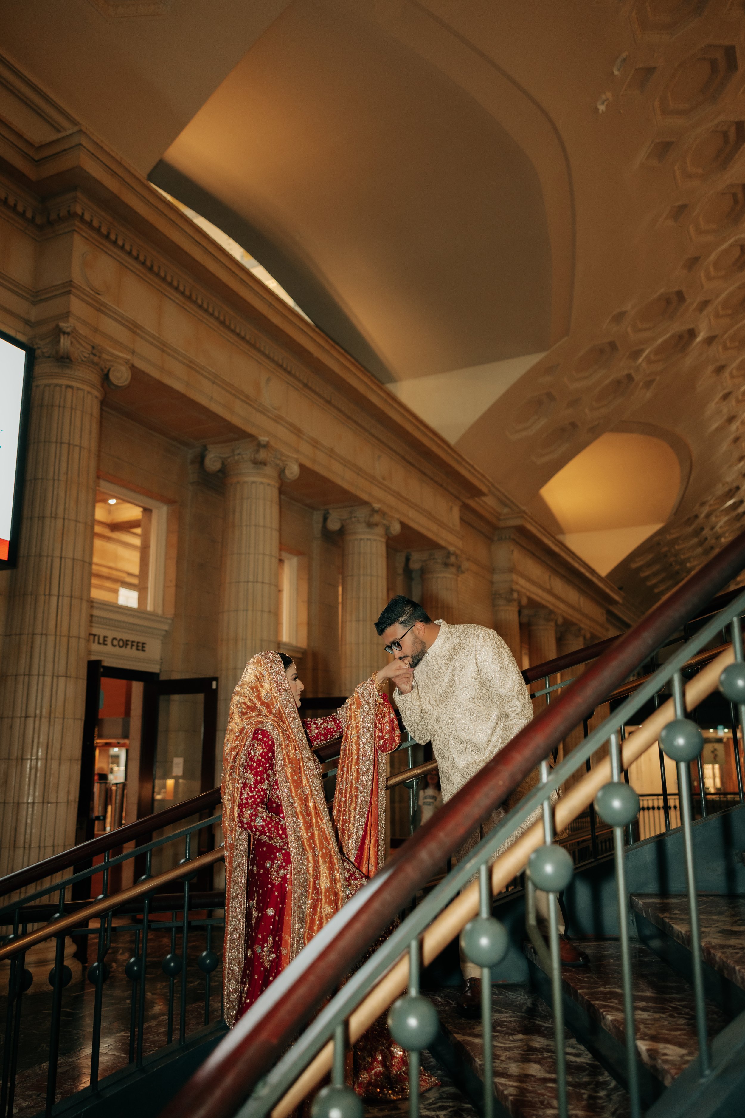 A couple dressed in traditional South Asian wedding attire standing on a staircase in an elegant, ornate building with classical architectural details.