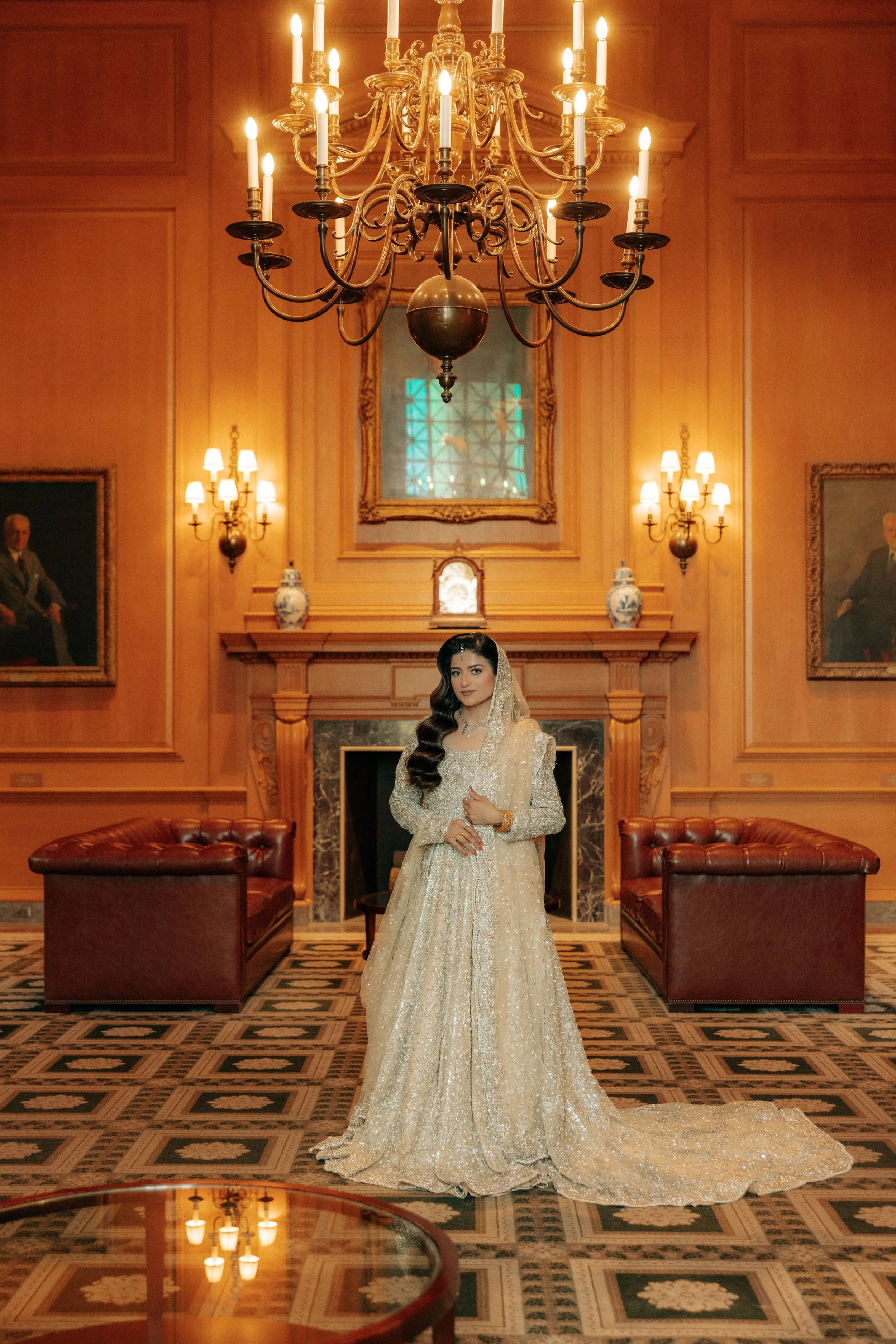 A woman in an elegant, cream-colored embellished gown stands in a lavish, wood-paneled room with a large chandelier hanging from the ceiling. The room features framed portraits on the walls, ornate vases on the mantel, and two leather sofas at the sides. The floor is decorated with a patterned carpet.