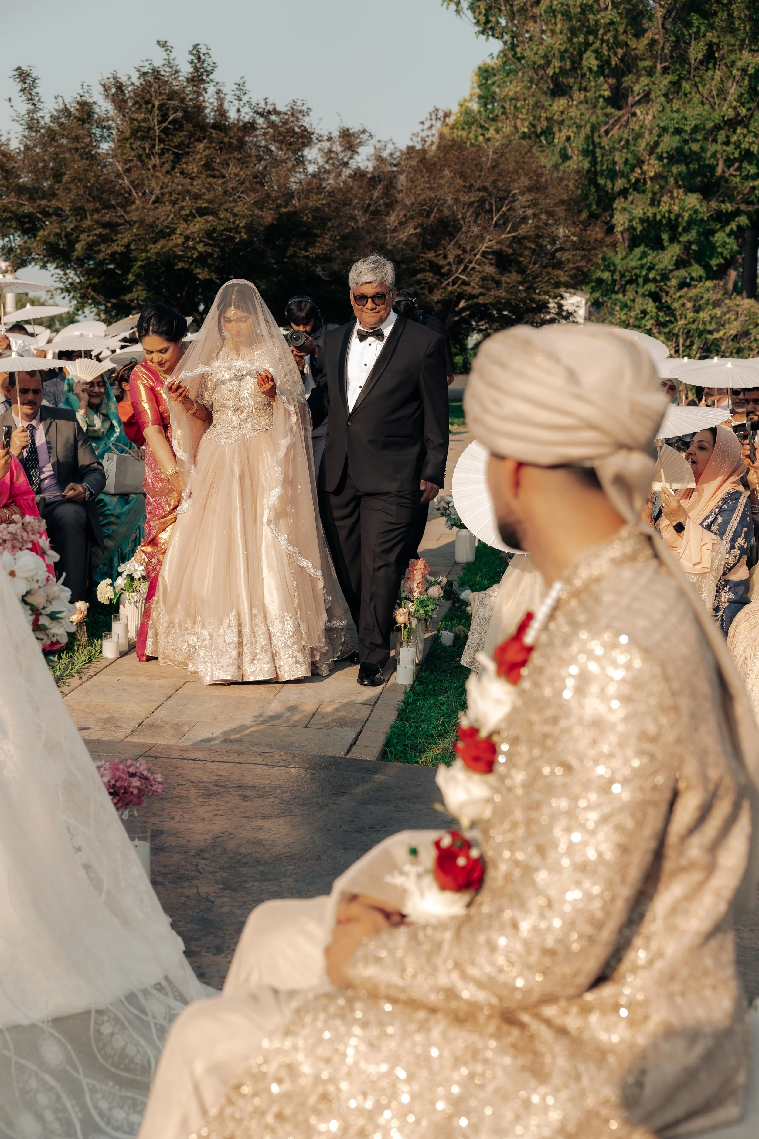 A wedding ceremony outdoors with bride and groom walking down the aisle, guests seated with white parasols, and formal attire including traditional clothing. Trees are visible in the background.