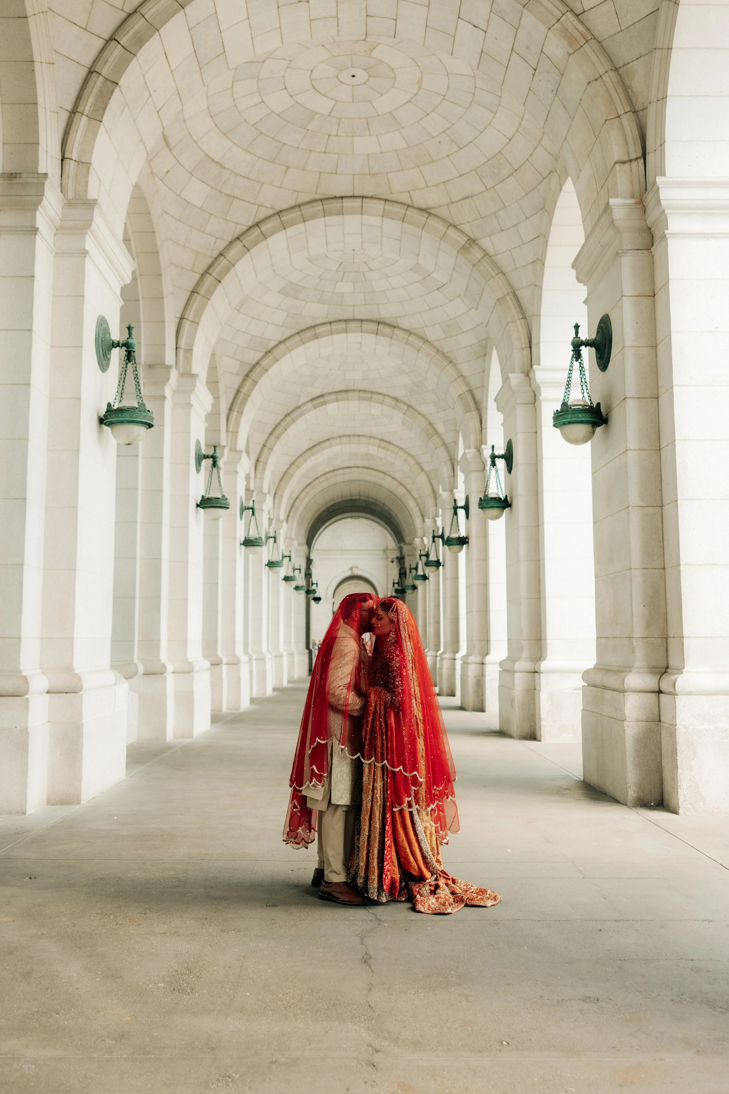 A couple dressed in traditional Indian wedding attire sharing a moment in an elegant stone archway with hanging lanterns.