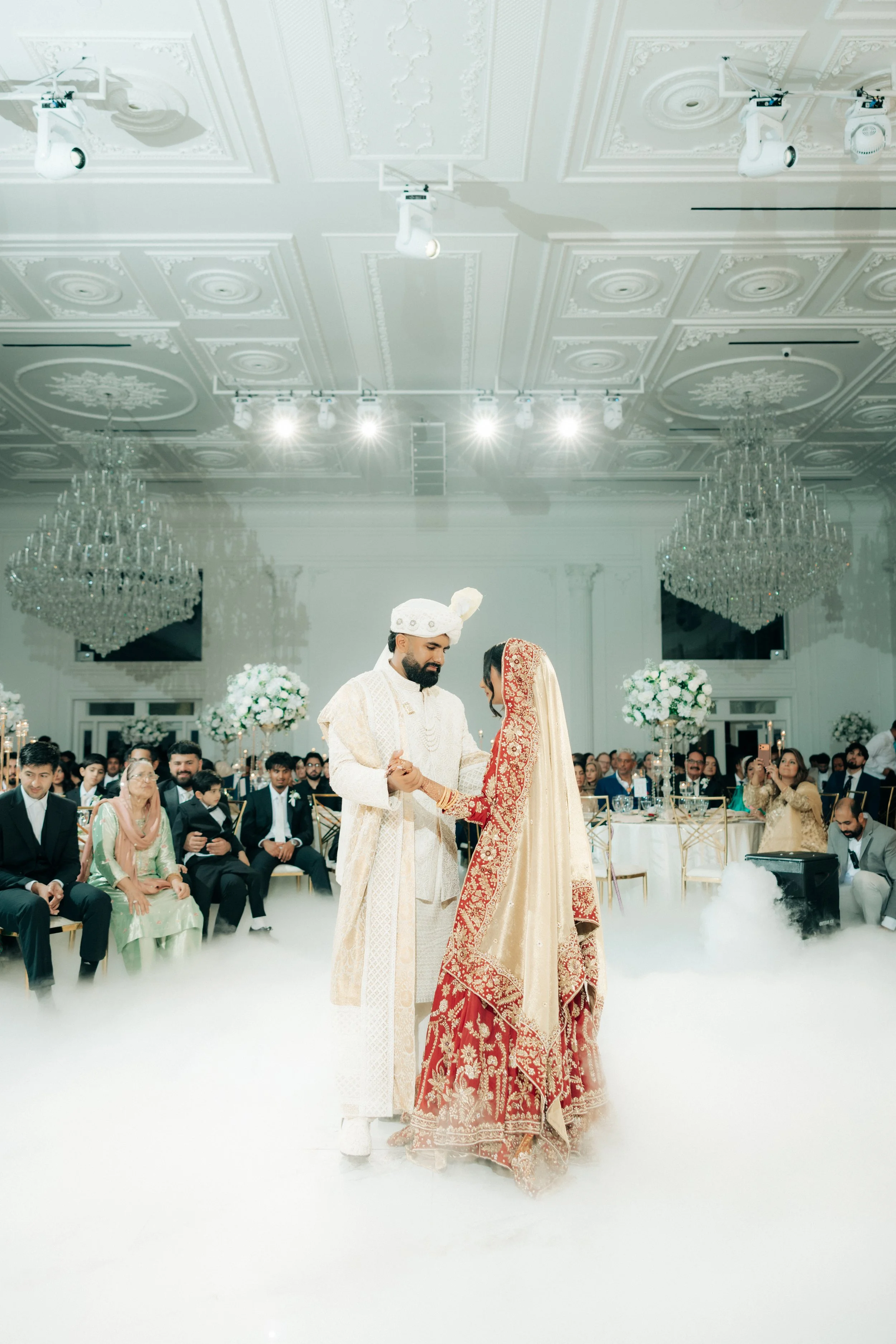 A traditional wedding ceremony with a couple dressed in ethnic wedding attire, holding hands and exchanging vows, surrounded by seated guests in an elegant, ornately decorated banquet hall with chandeliers and floral arrangements.