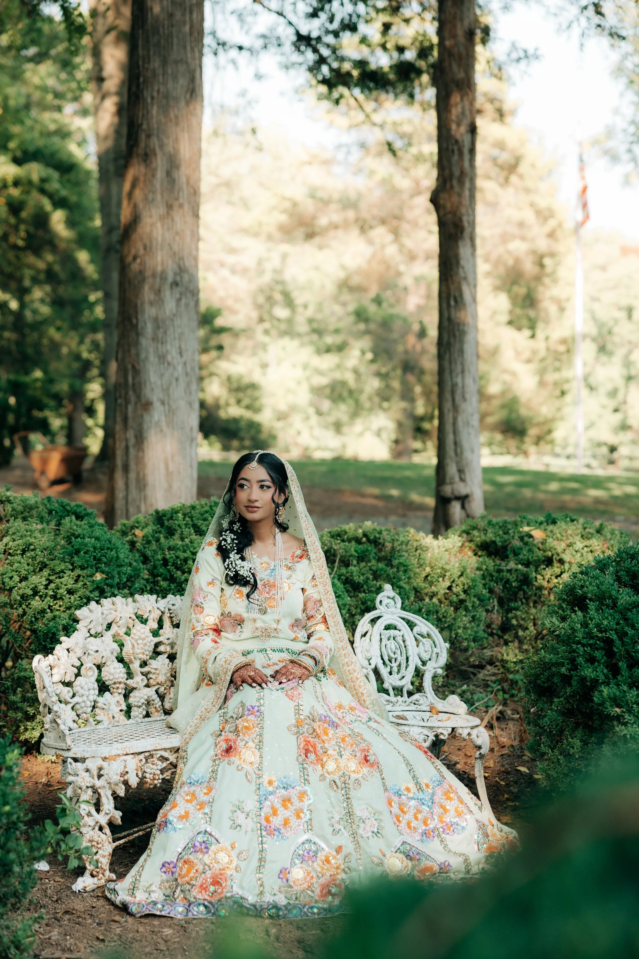 A young woman in a traditional embroidered dress with floral patterns and henna on her hands, sitting on an ornate white garden bench in a lush, sunlit park with tall trees and green bushes in the background.