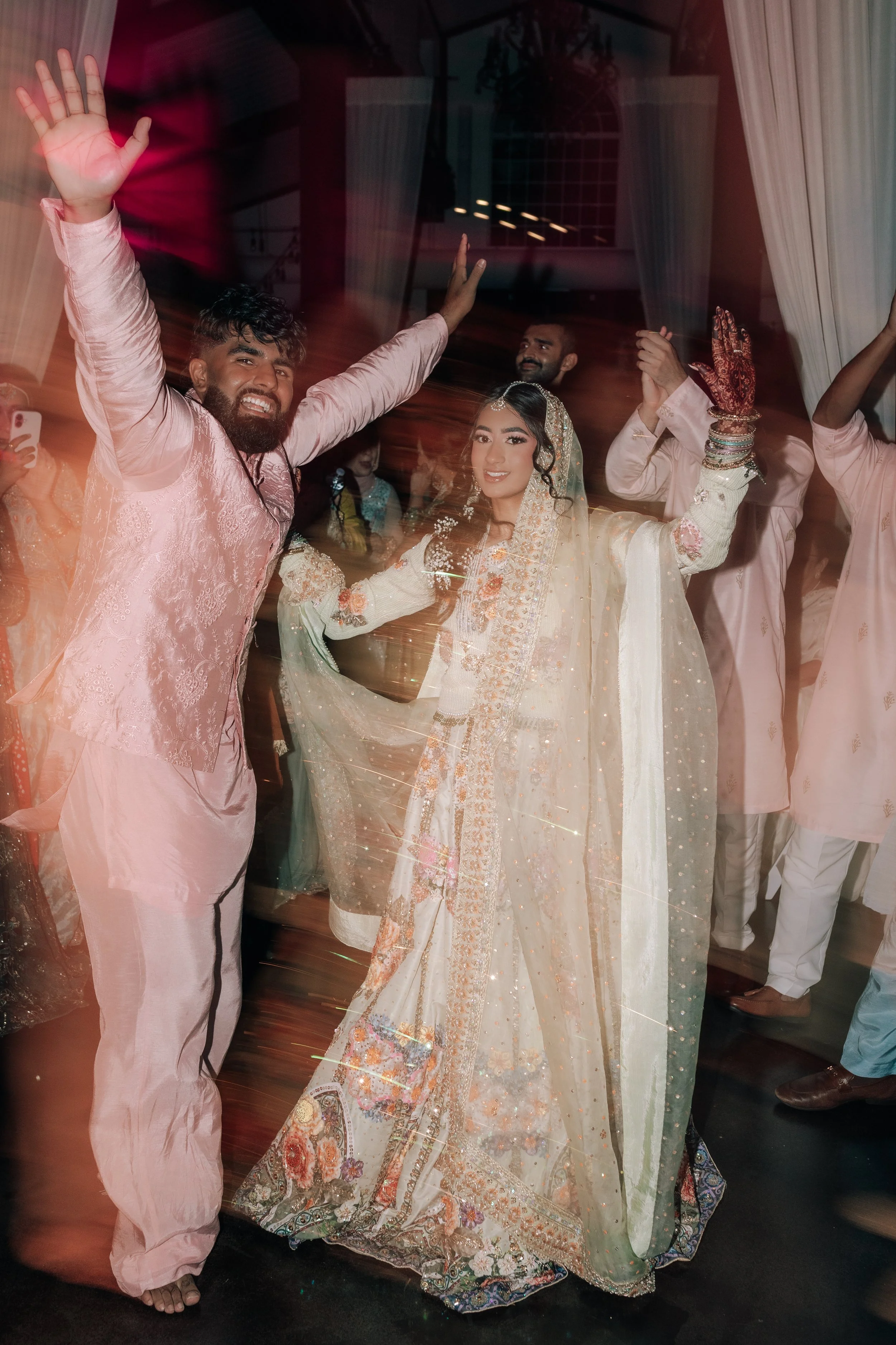 A woman in traditional South Asian attire, likely at a celebration or wedding, is dancing with her arms raised. She is surrounded by men dressed in similar attire, with some raising their hands in celebration. The scene is indoors with curtains and a