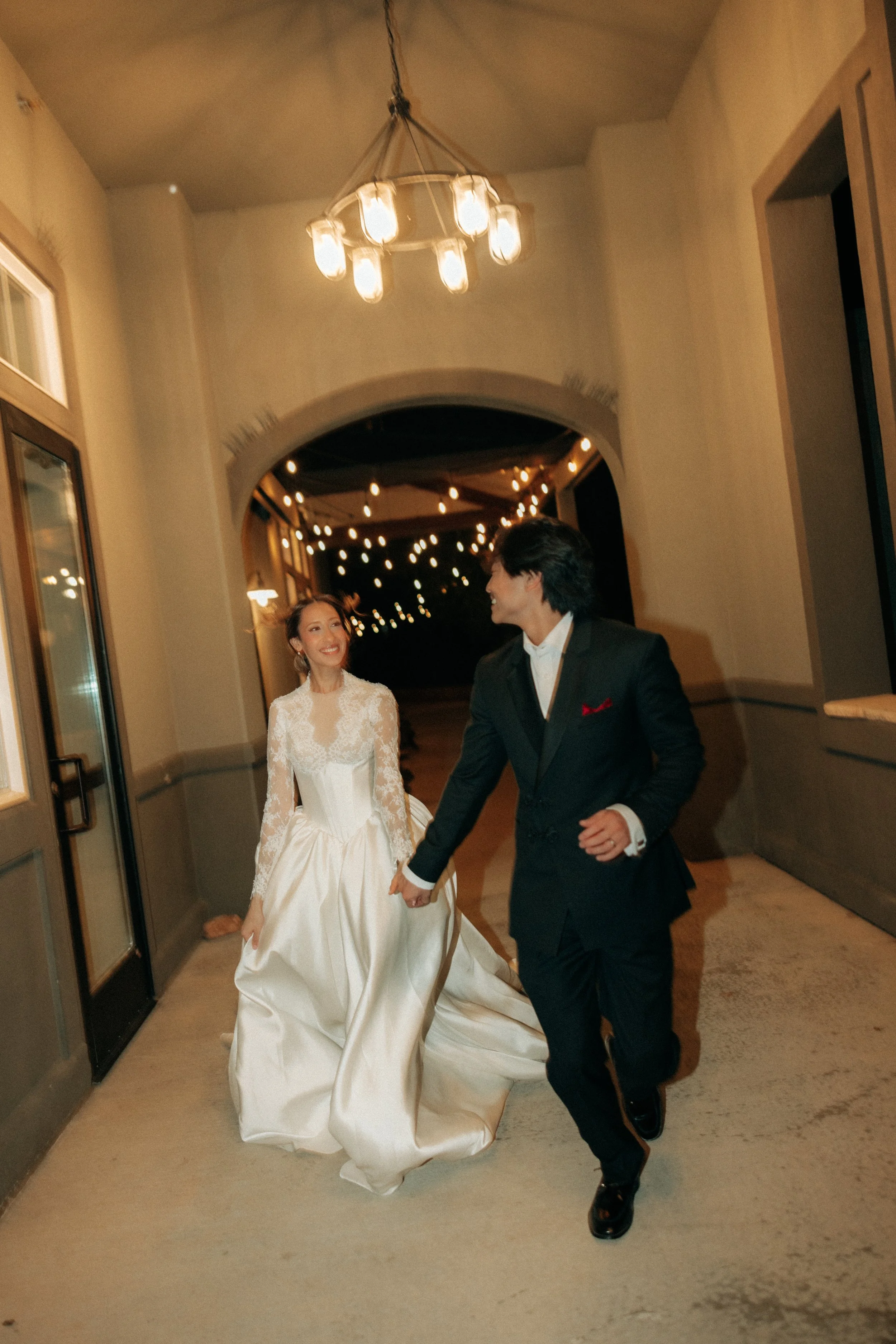 A bride and groom walking hand in hand through a hallway at night. The bride is wearing a white wedding gown with lace and a full skirt, while the groom is dressed in a black tuxedo with a white shirt and red pocket square. They are smiling at each o