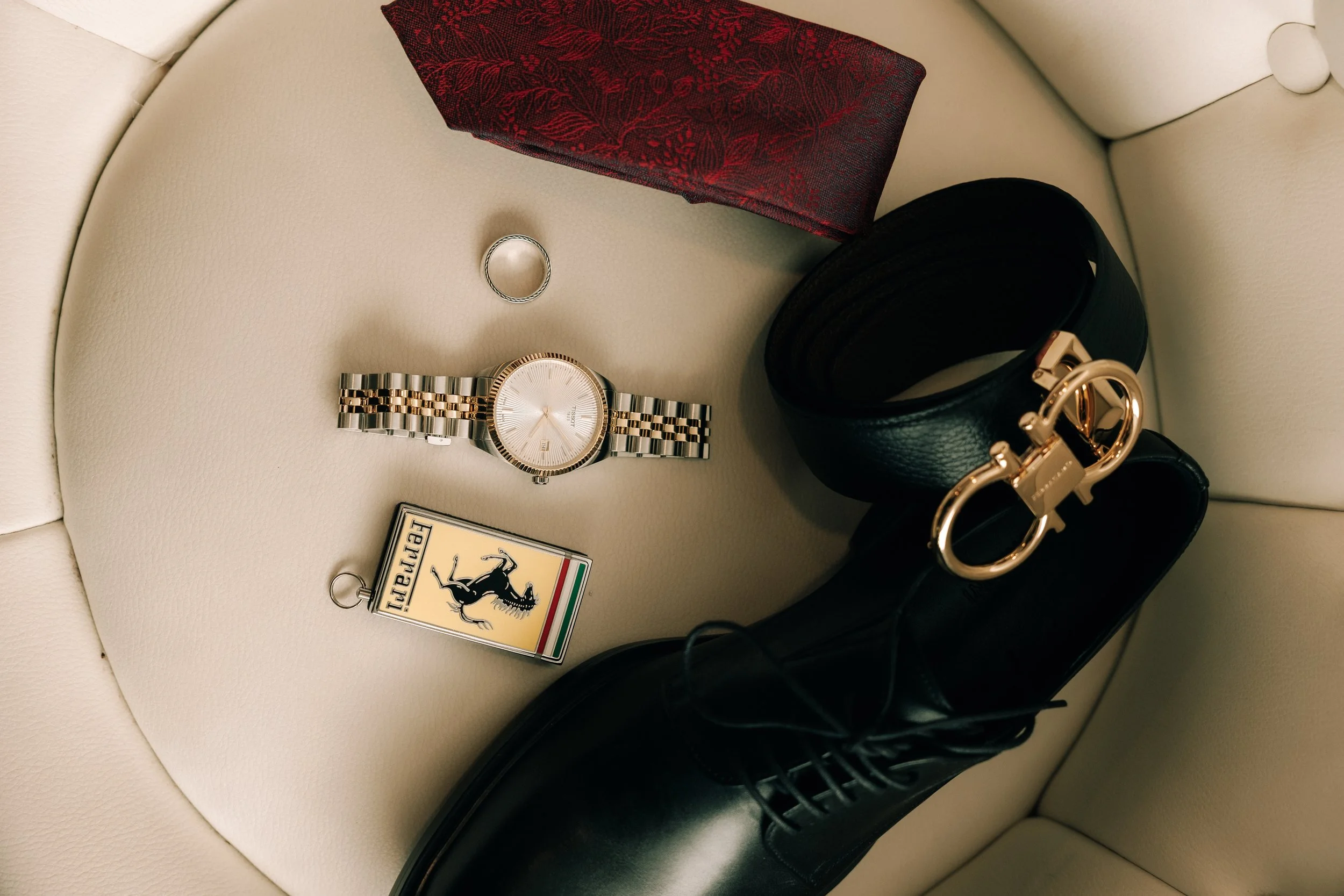 A beige curved leather surface with a wristwatch, a ring, a red fabric item, black belt, black shoes, a leather purse, a pack of cigarettes, and a keychain with a Ferrari logo.