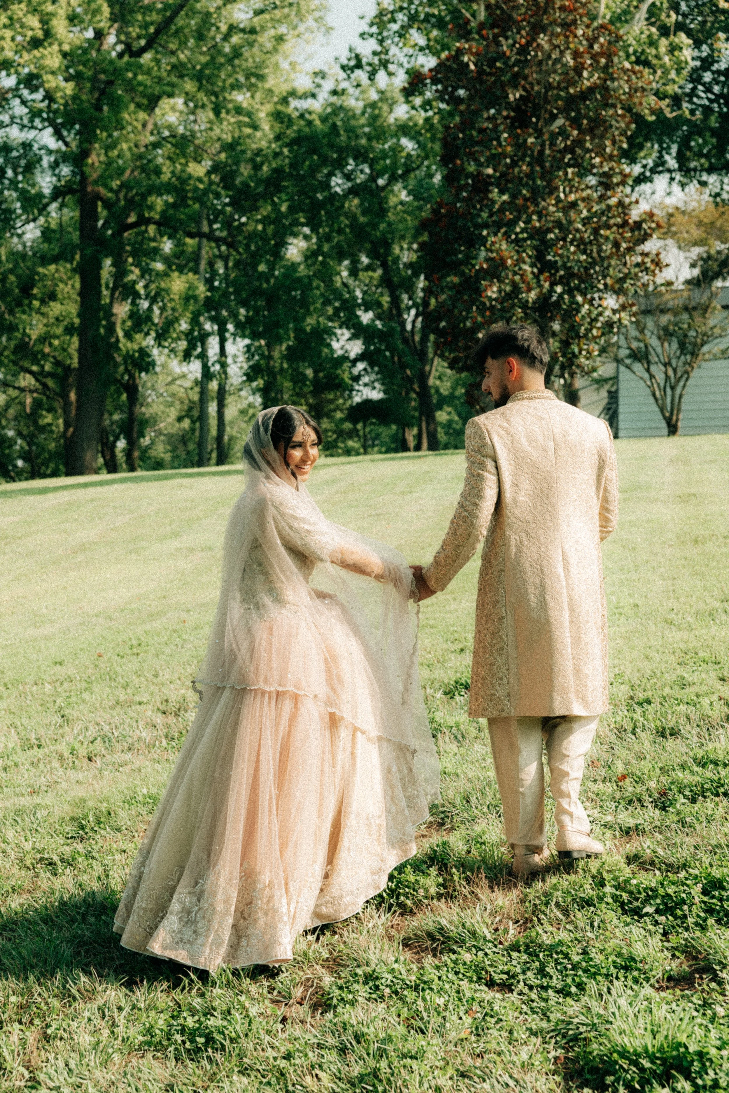 A couple dressed in traditional, elegant wedding attire holding hands in a green outdoor park with trees and a house in the background.
