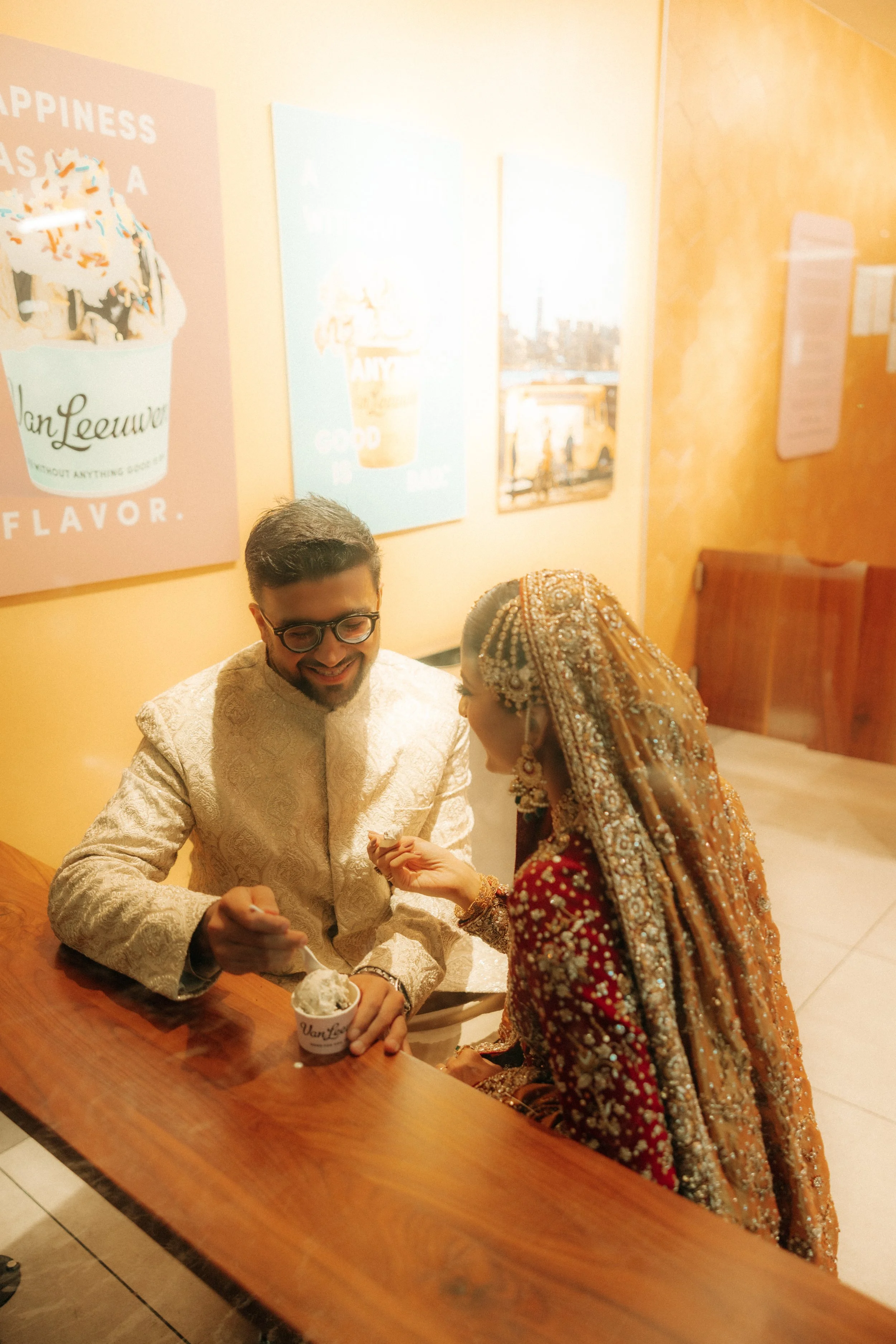 A couple dressed in traditional Indian wedding attire sharing a moment while eating ice cream at a wooden table in a brightly lit room with yellow walls and colorful wall art.