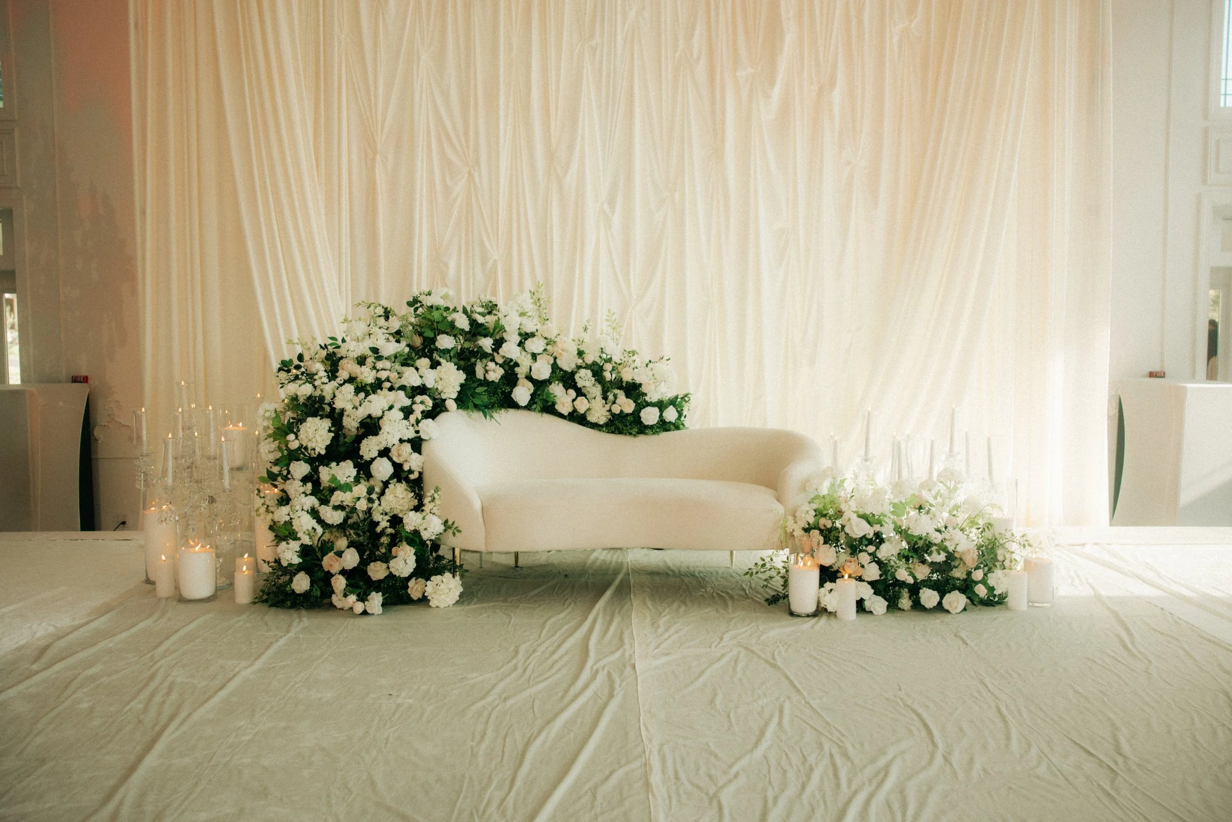 Elegant floral wedding altar with a white loveseat, surrounded by white flowers and candles, set against a cream-colored curtain backdrop.