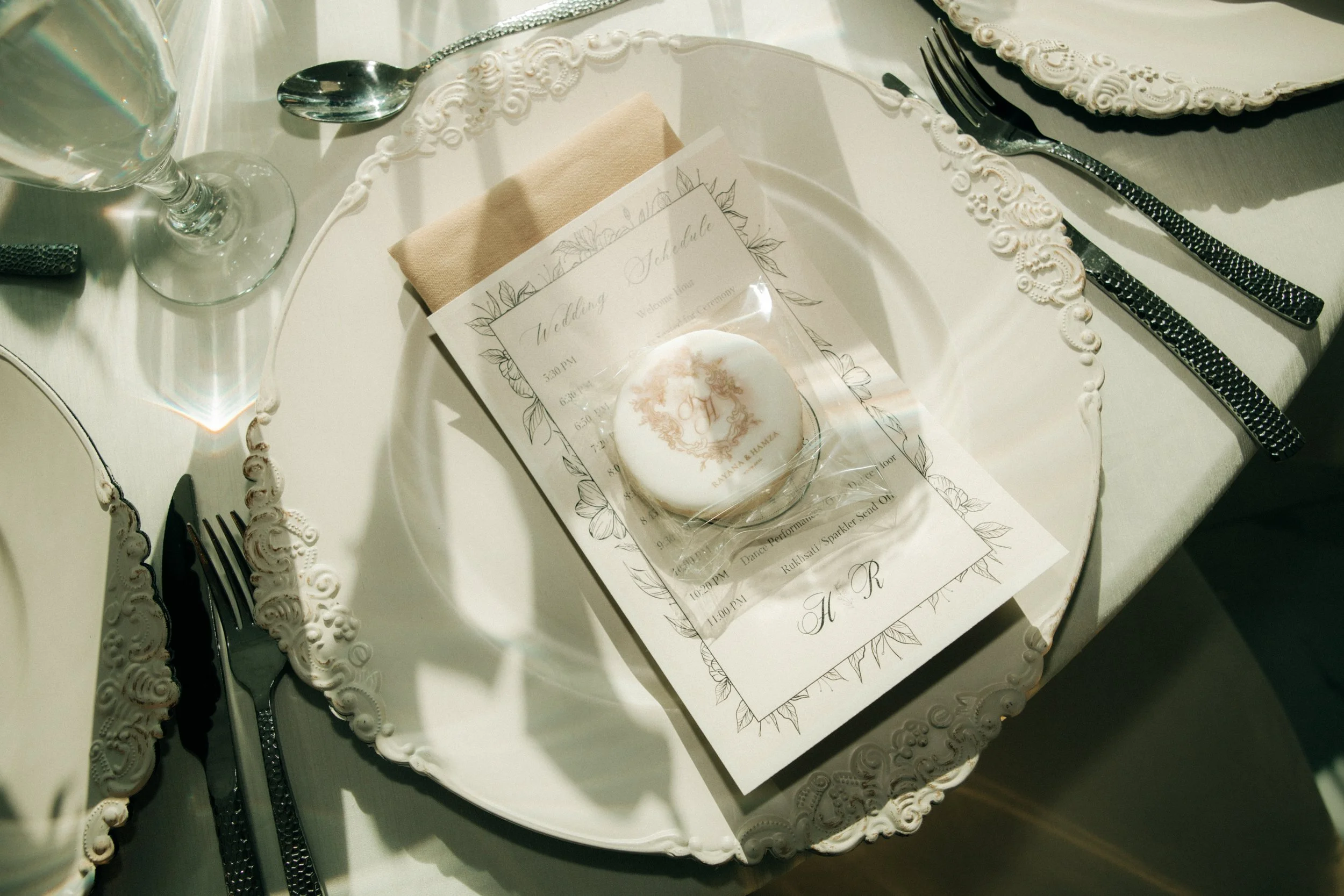 Set of ornate white serving trays with intricate designs, set on a table with cutlery, a glass of water, a wrapped favor, and wedding schedule cards.