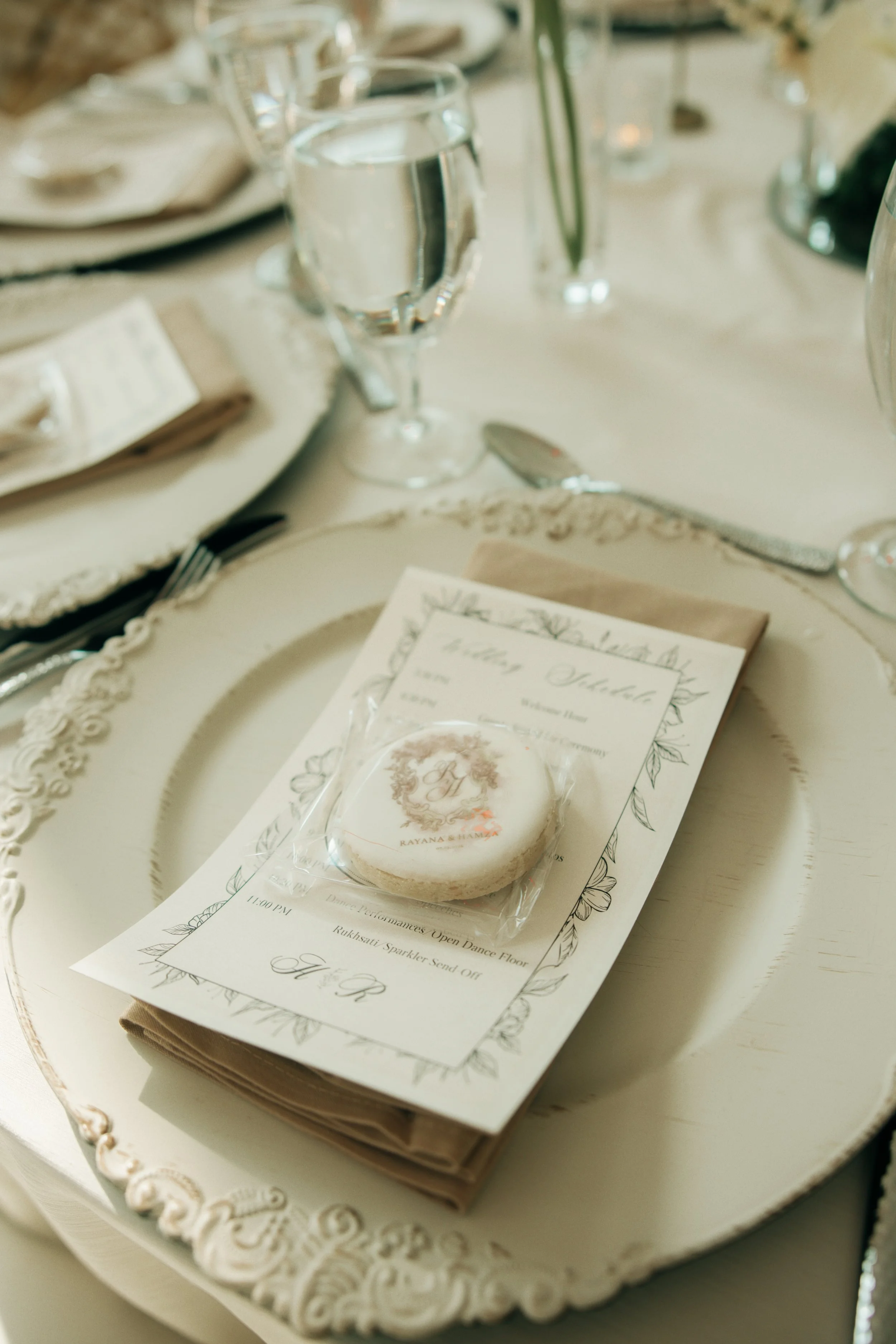 A wedding invitation card sitting on a table with a small round decorative cake on top. The table is set with glasses of water, silverware, and plates. The setup is elegant with floral-themed decor.