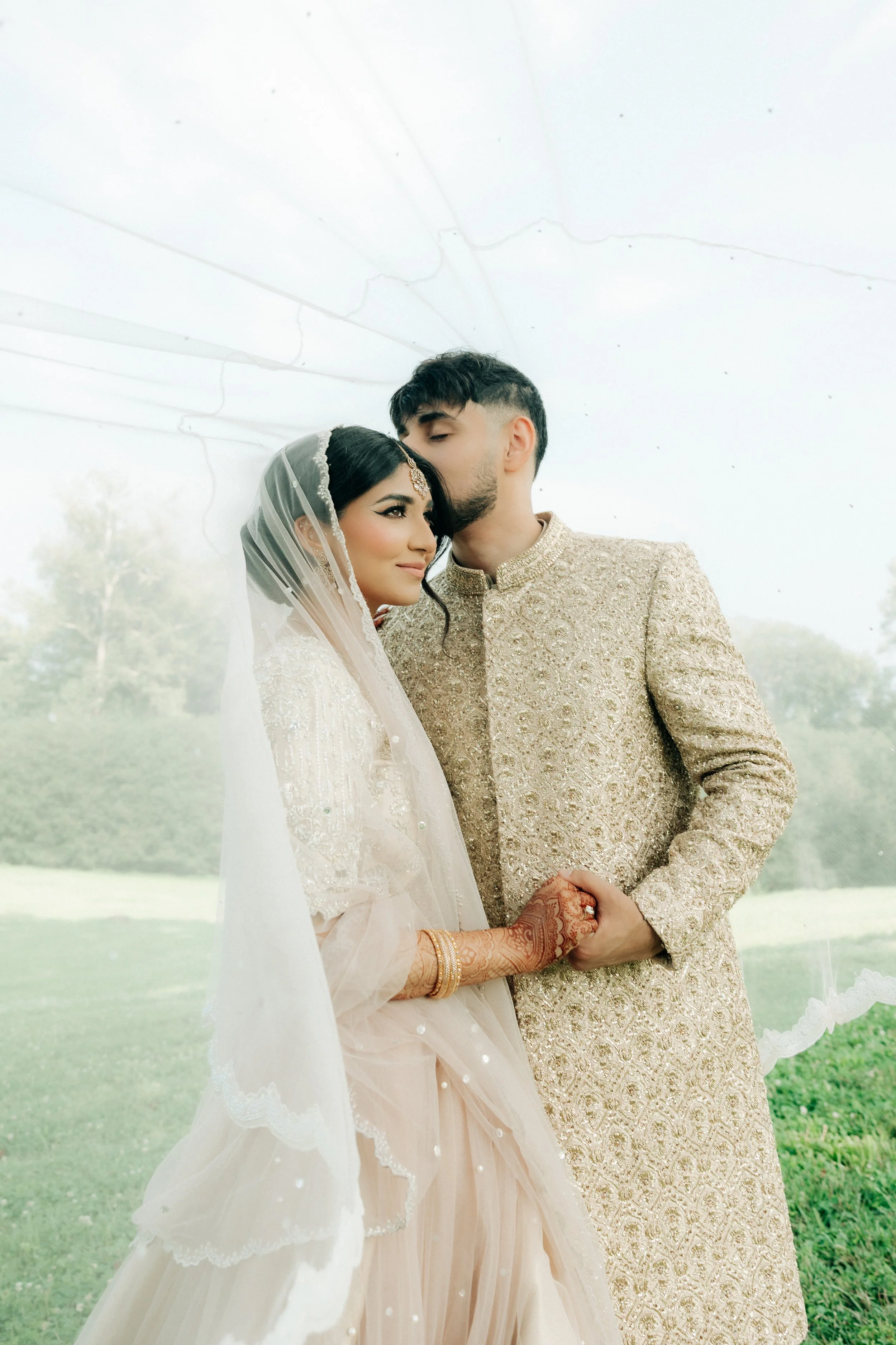 A bride and groom holding hands, with the groom leaning in to kiss the bride on the forehead, outdoors with a grassy background.