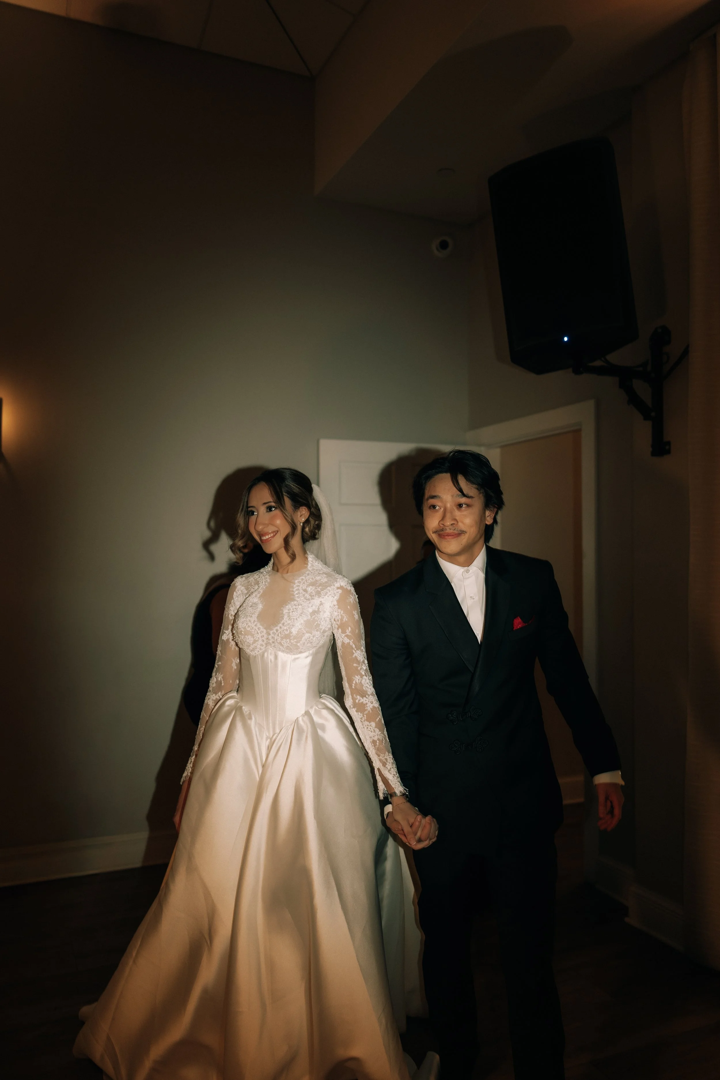 A bride and groom holding hands at their wedding reception, smiling warmly. The bride is wearing a cream lace wedding dress, and the groom is dressed in a black suit and white shirt. They are standing indoors, against a plain wall with a speaker moun