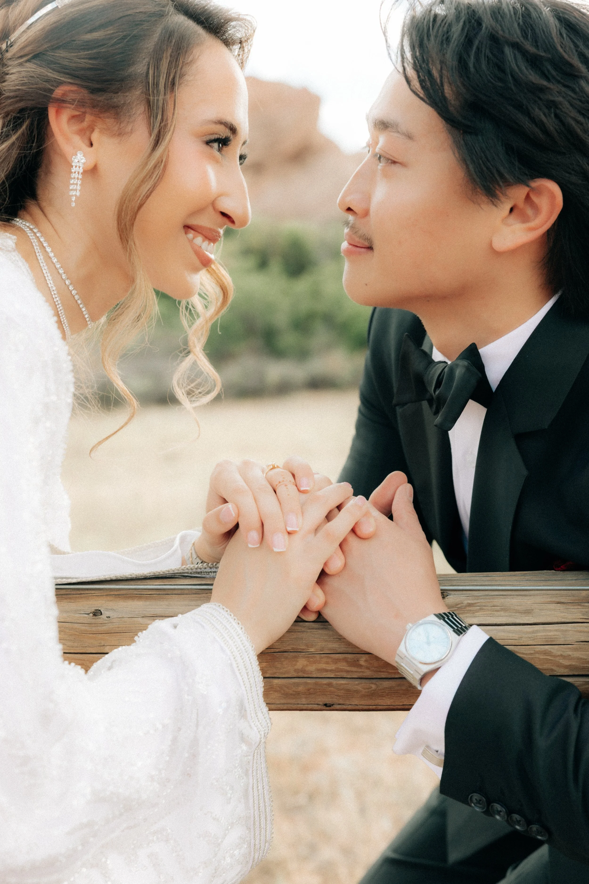 A bride and groom sitting at a wooden table face to face, holding hands, outdoors with a desert landscape in the background. The bride has wavy hair, wears pearl earrings and necklace, and a white dress. The groom wears a black tuxedo with a bow tie,
