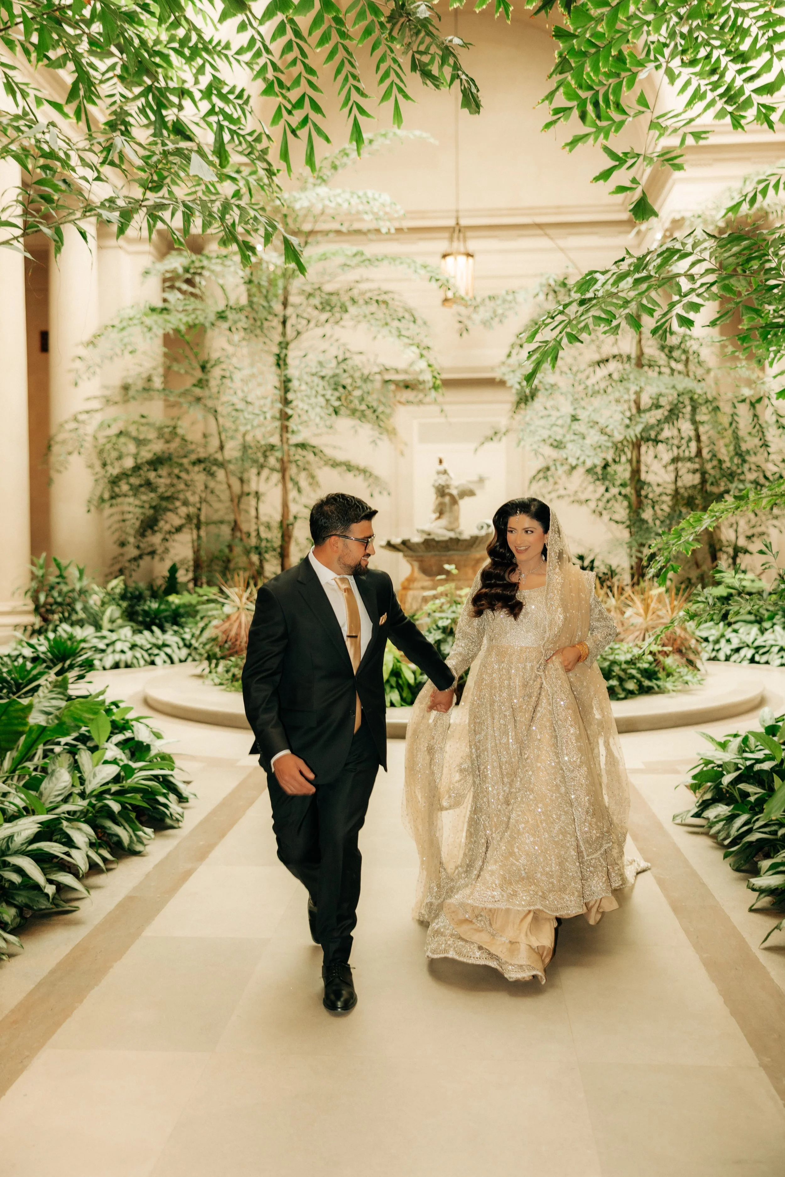 A bride and groom walking hand in hand in an elegant indoor setting filled with lush green plants and trees, with a fountain in the background.