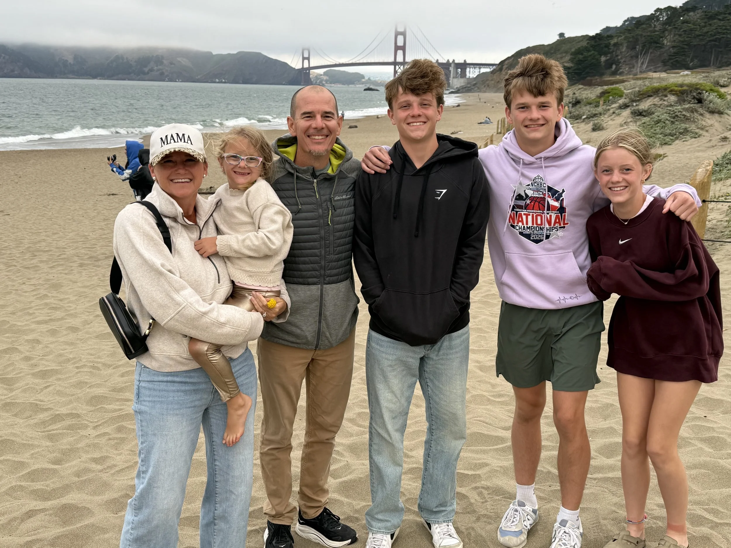 A group of seven people, including children and adults, smiling and standing on a sandy beach with the Golden Gate Bridge in the background. It is cloudy and overcast.