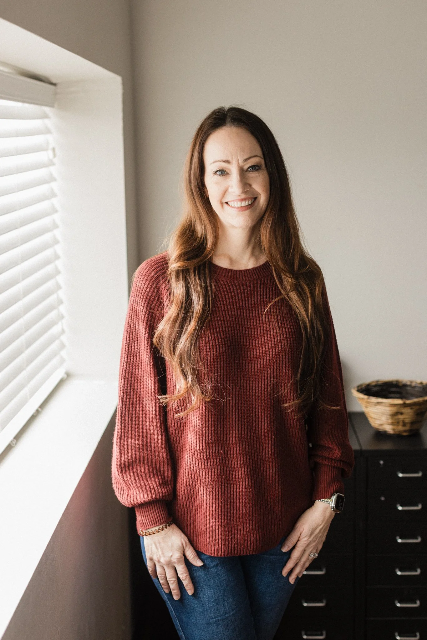 A woman with long, wavy brown hair and light skin, smiling, wearing a red sweater, standing indoors near white blinds and a dark drawer.