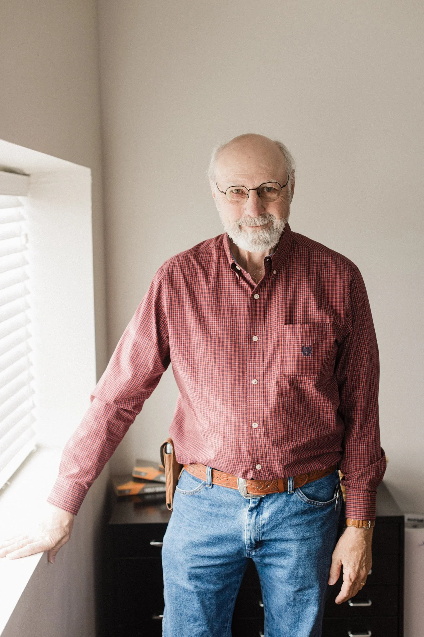 An older man wearing glasses, a red plaid shirt, jeans, and a leather belt, standing indoors next to a window with white blinds, with a drawer visible behind him.