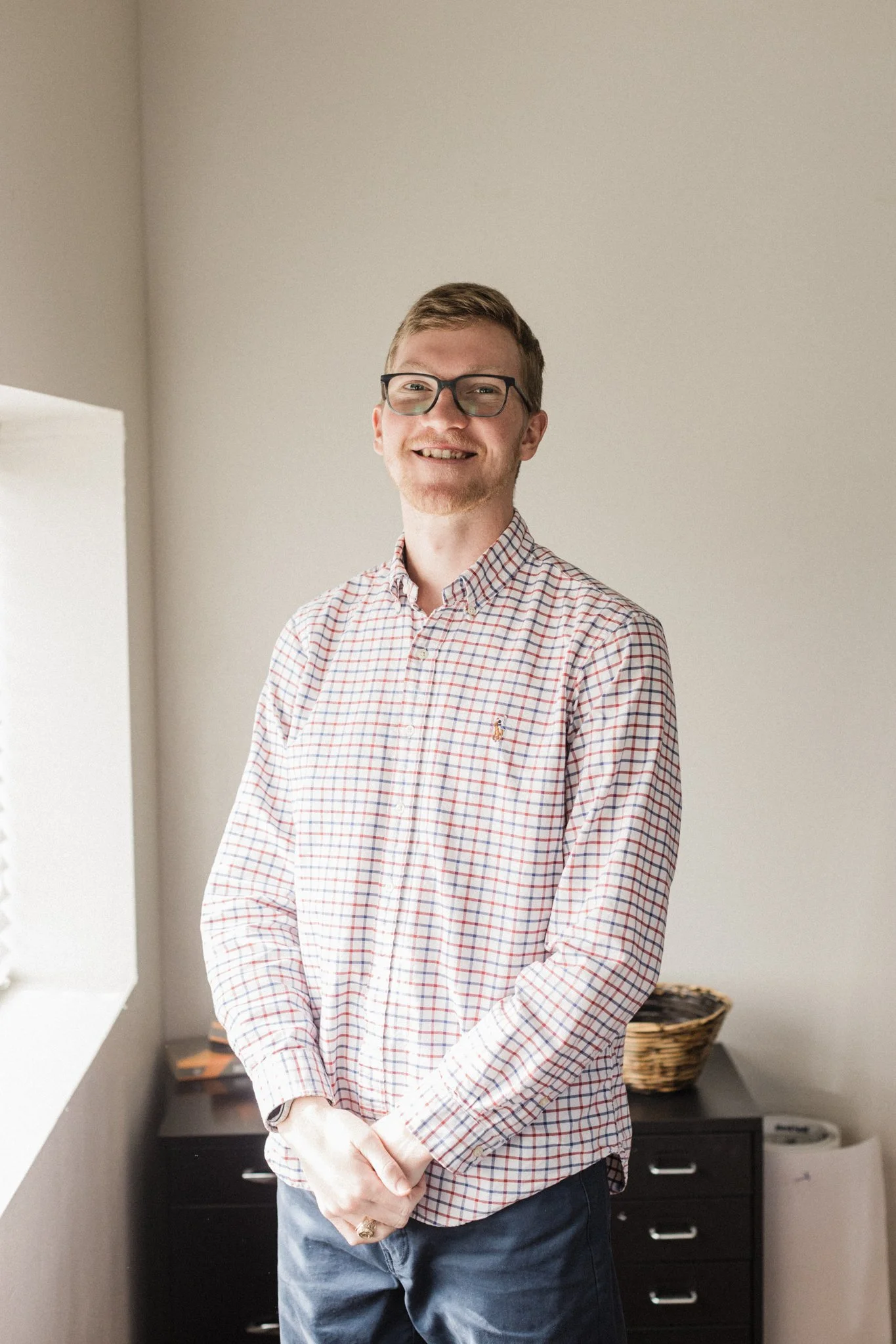 A young man with glasses and short hair smiling, wearing a checkered shirt and jeans, standing indoors near a window with a black desk and basket in the background.