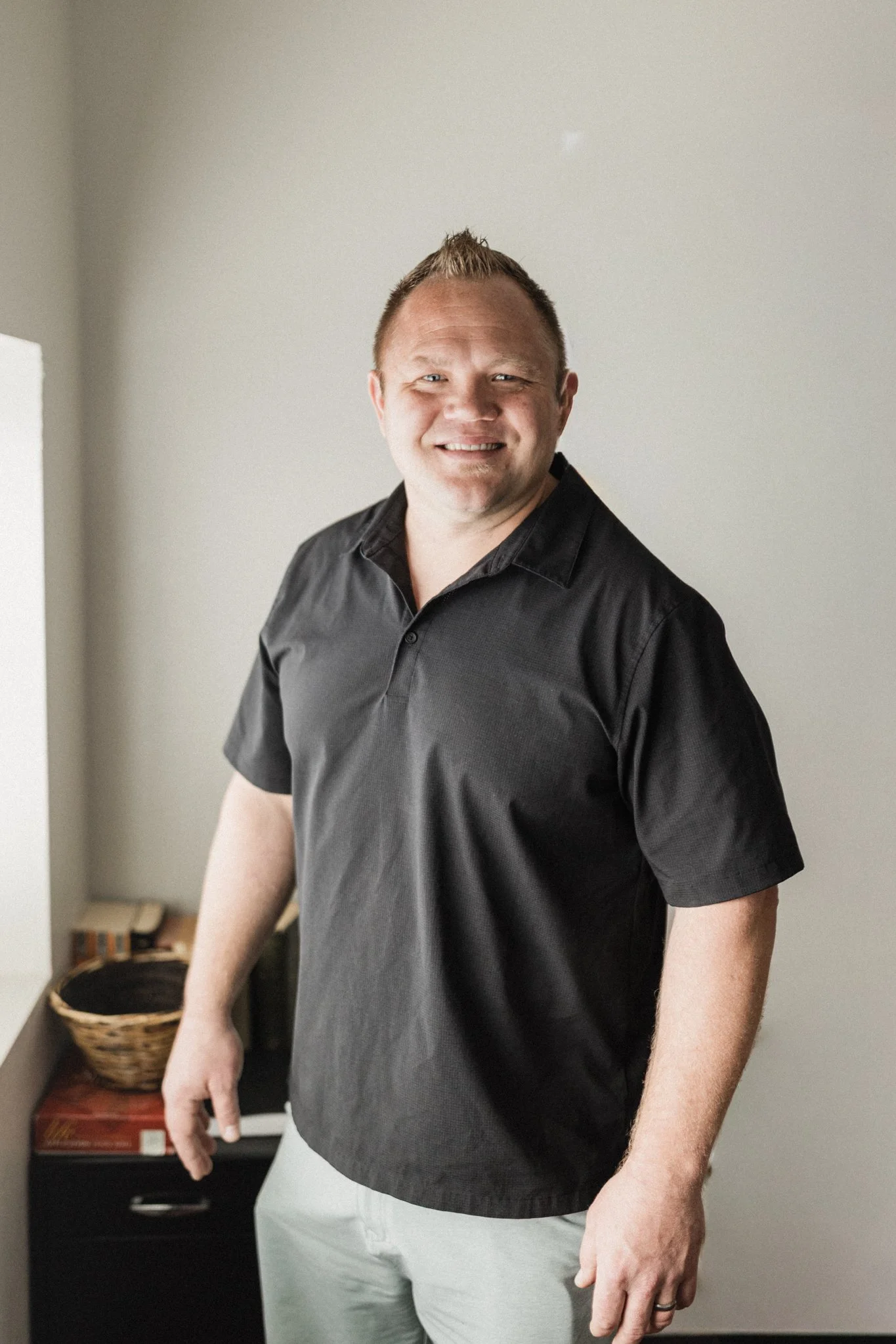 A smiling man with short light brown hair, wearing a black shirt and light-colored pants, standing inside a room with a basket and a table in the background.