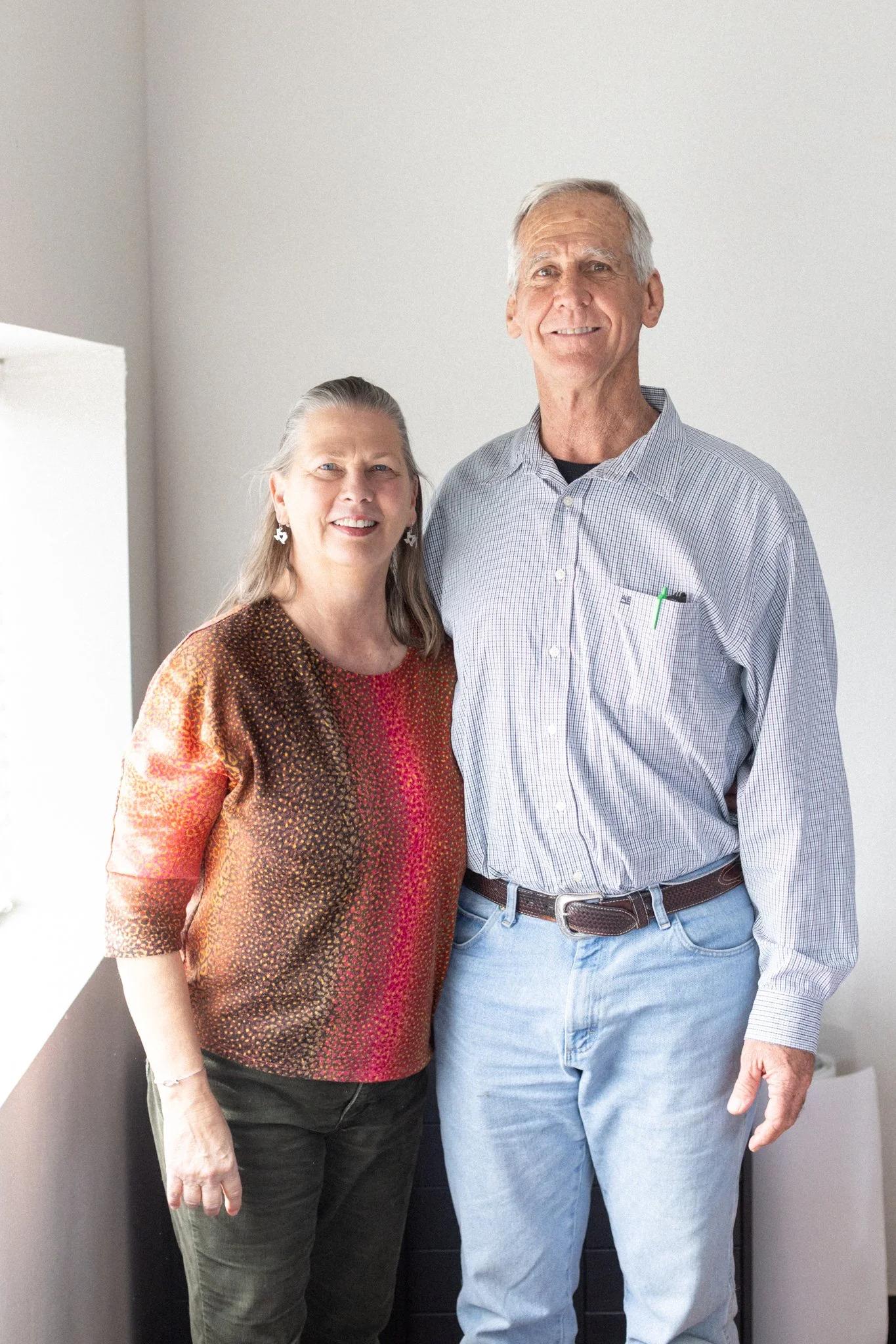 A middle-aged woman with long gray hair wearing a multicolored, patterned top standing next to an older man with short gray hair wearing a light blue checked shirt and light blue jeans, both smiling and looking at the camera.