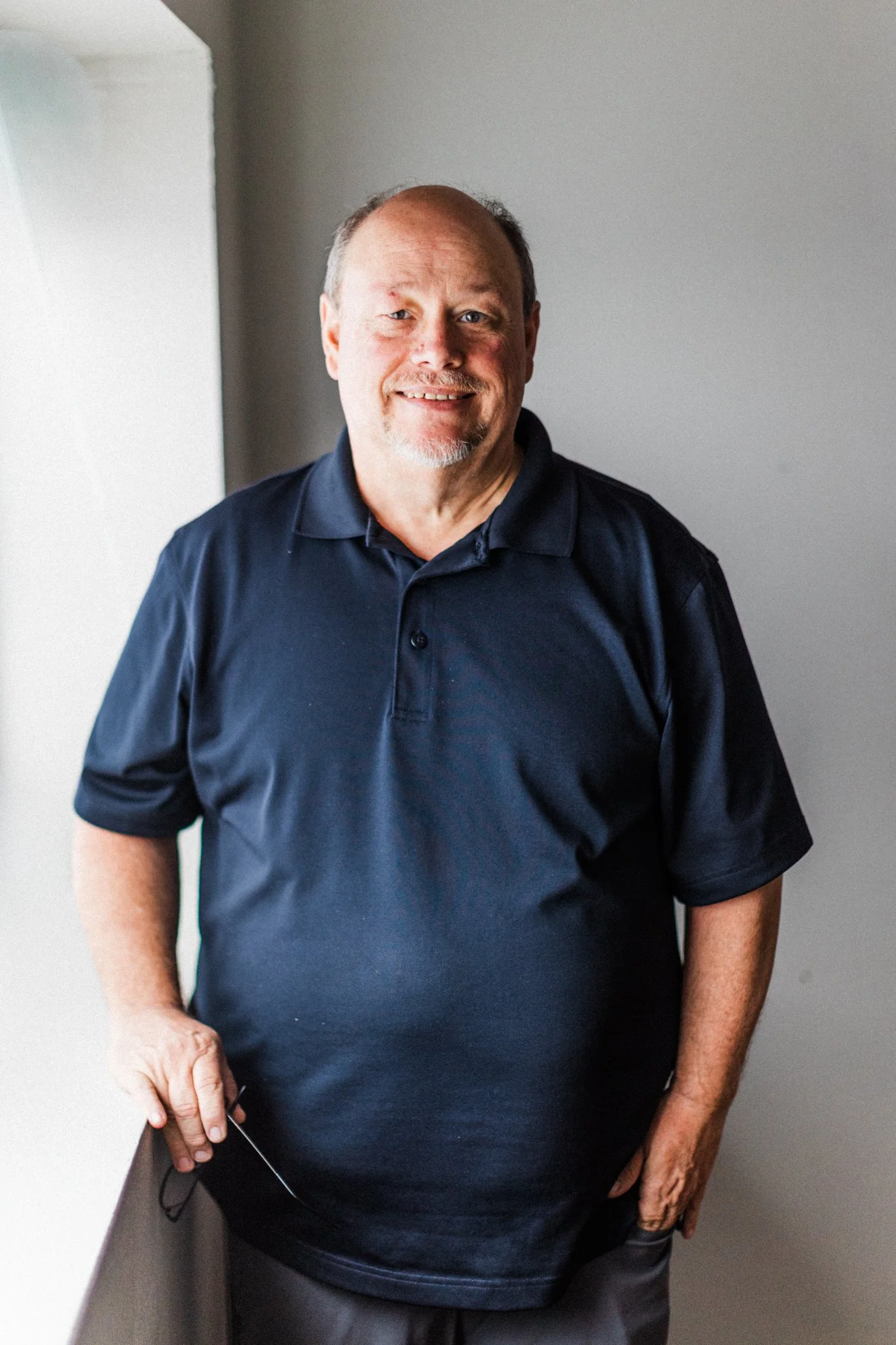 A middle-aged man with a goatee and receding hairline standing inside, smiling at the camera. He's wearing a navy blue polo shirt and dark pants, holding a pair of eyeglasses in his right hand, with a plain wall as the background.