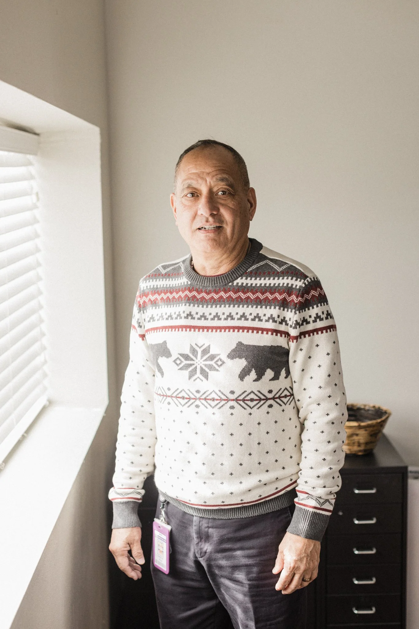 A man in a Christmas sweater with polar bears and snowflakes standing indoors near a window with white blinds.