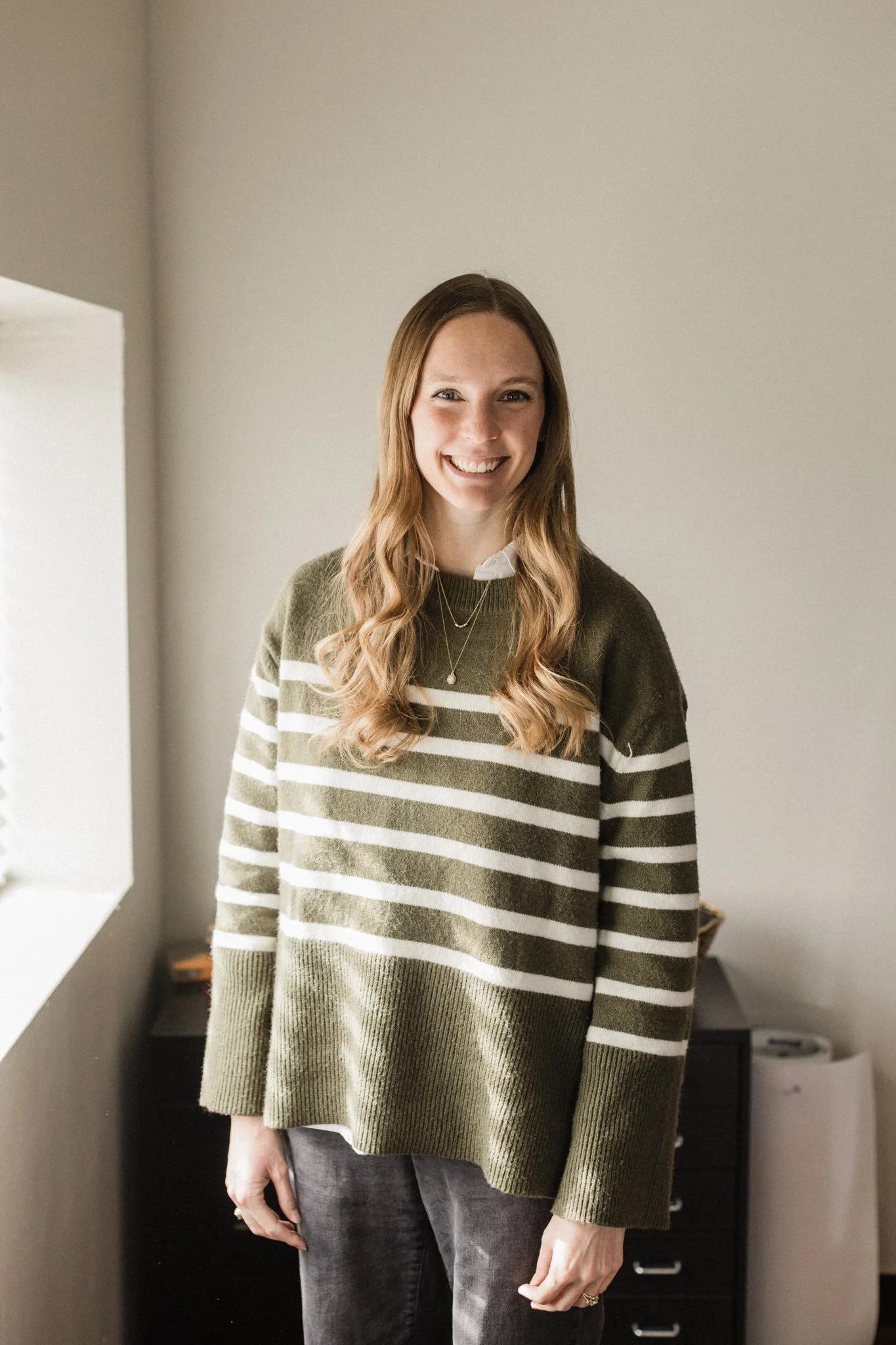 A woman with long wavy hair smiling, wearing a green and white striped sweater, gray jeans, and a necklace, standing inside a room with a neutral-colored wall.